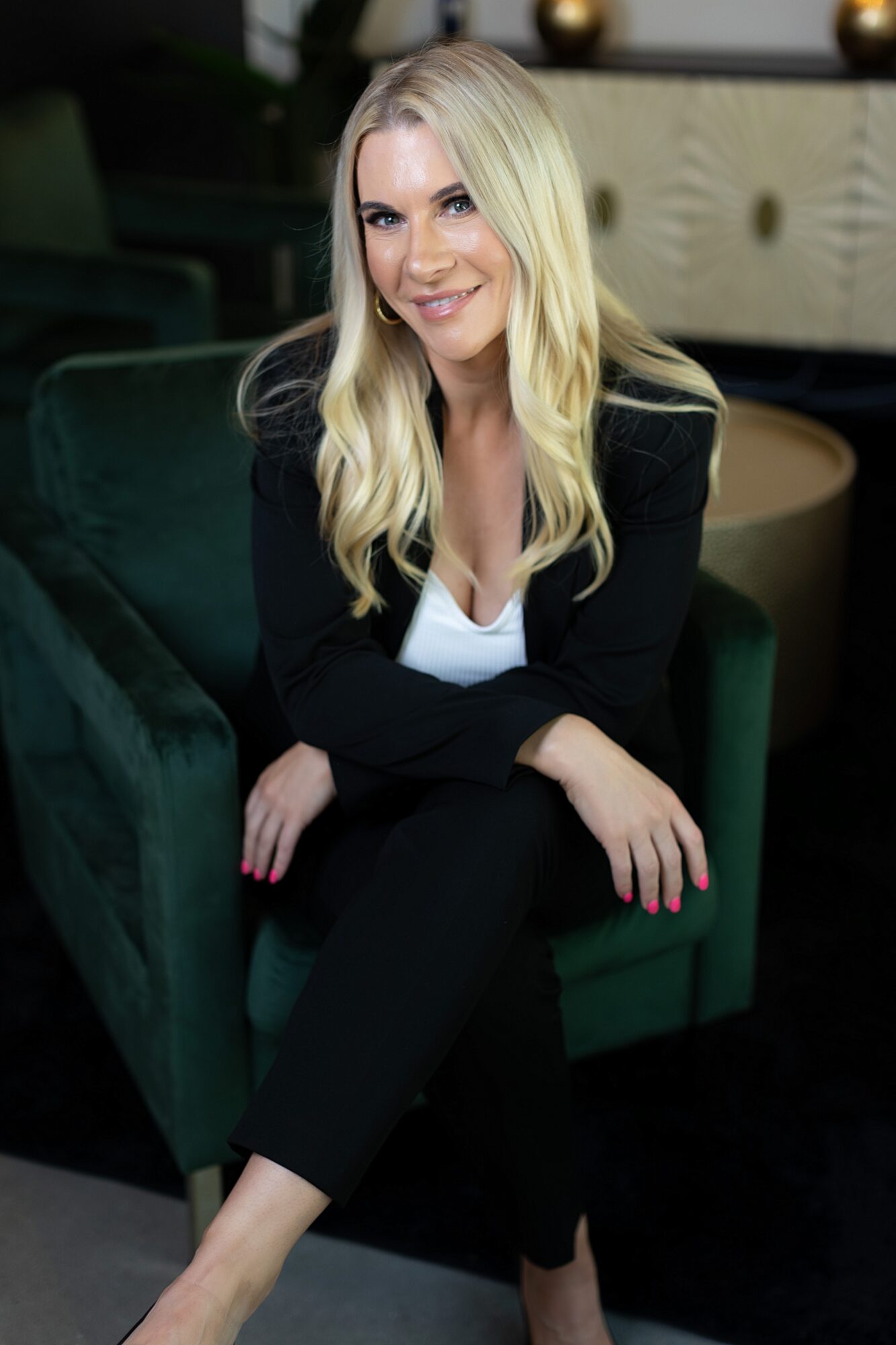Woman with long blonde hair smiling, sitting on a green armchair in a modern interior, wearing a black blazer and white top.