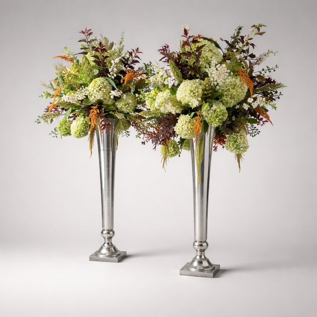 Two tall silver vases with floral arrangements of green, white, and orange flowers and foliage, on a plain background.