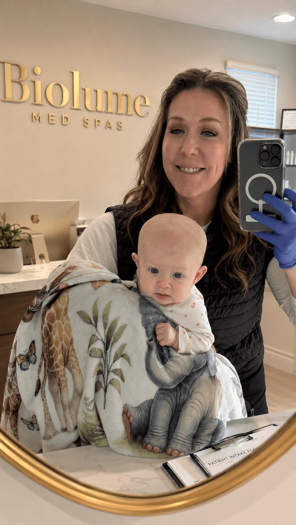 Woman holding a baby in a spa reception area, smiling, with a sign reading 'Biolume' in the background.
