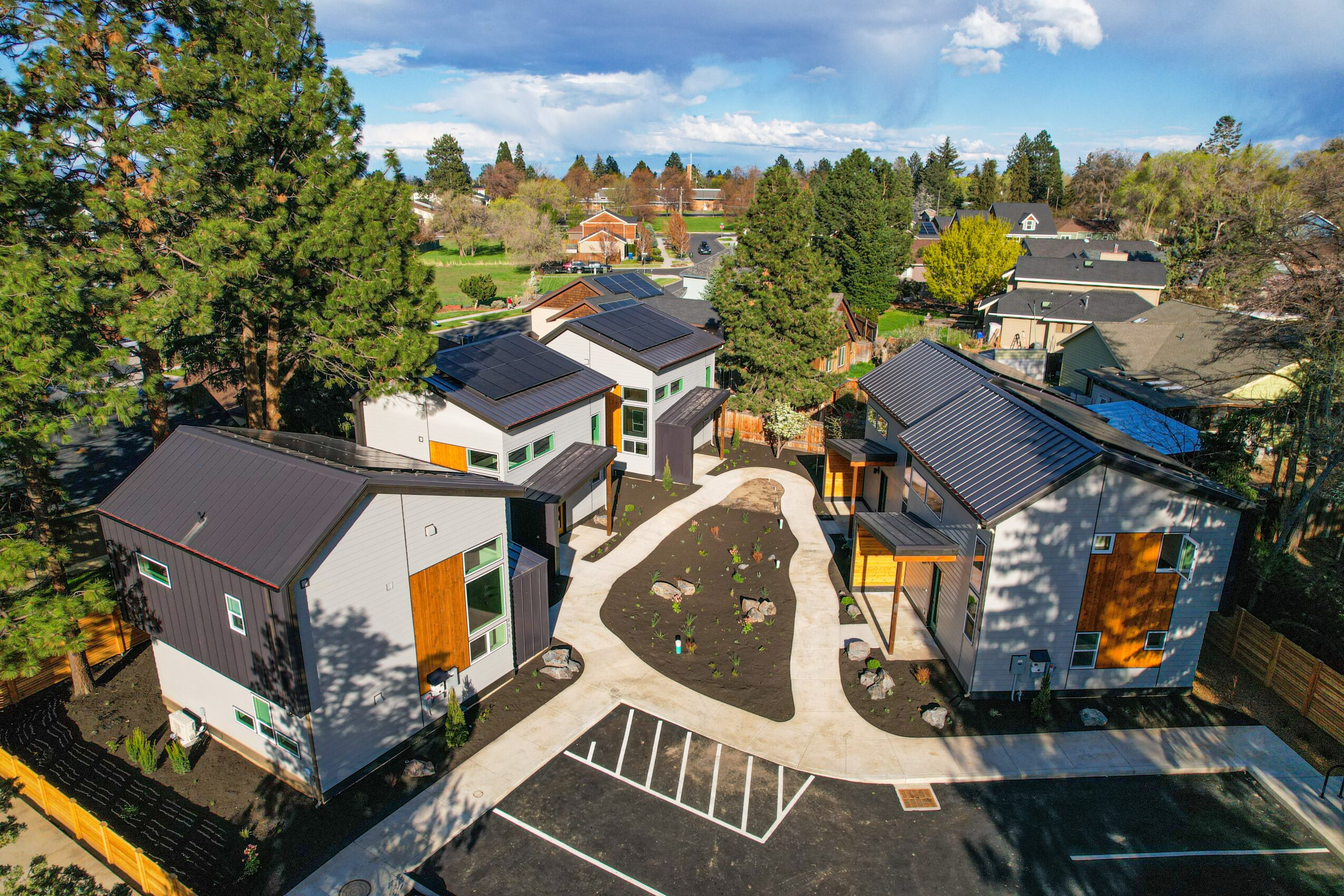 Residential complex with modern houses, trees, and a parking area under a partly cloudy sky.