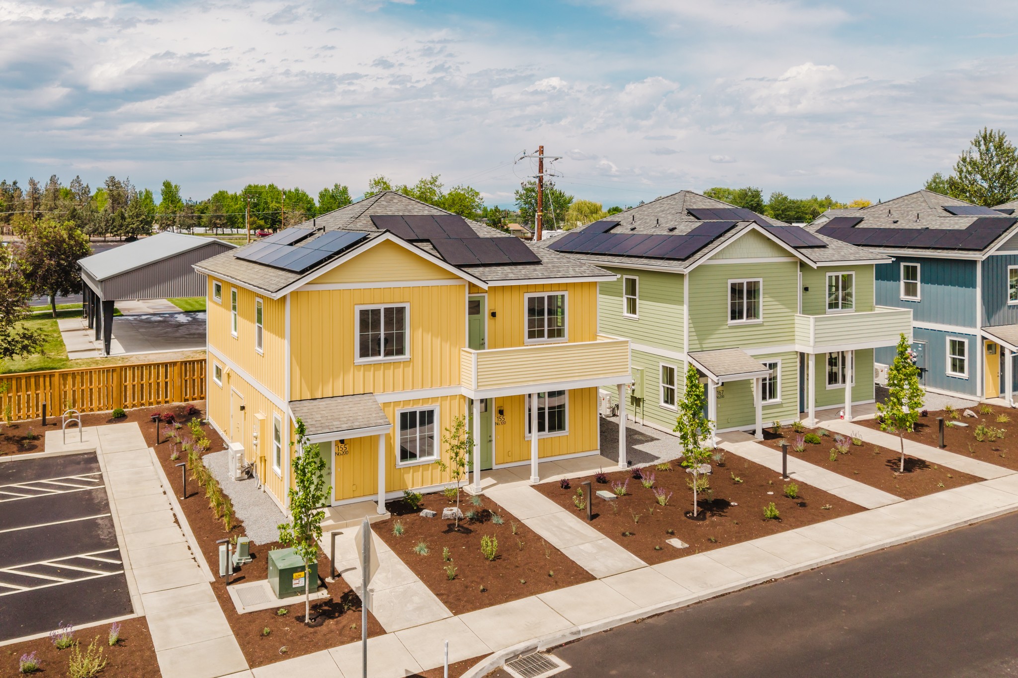 Colorful row of new houses with small front yards, trees, and parking lot, under a partly cloudy sky.