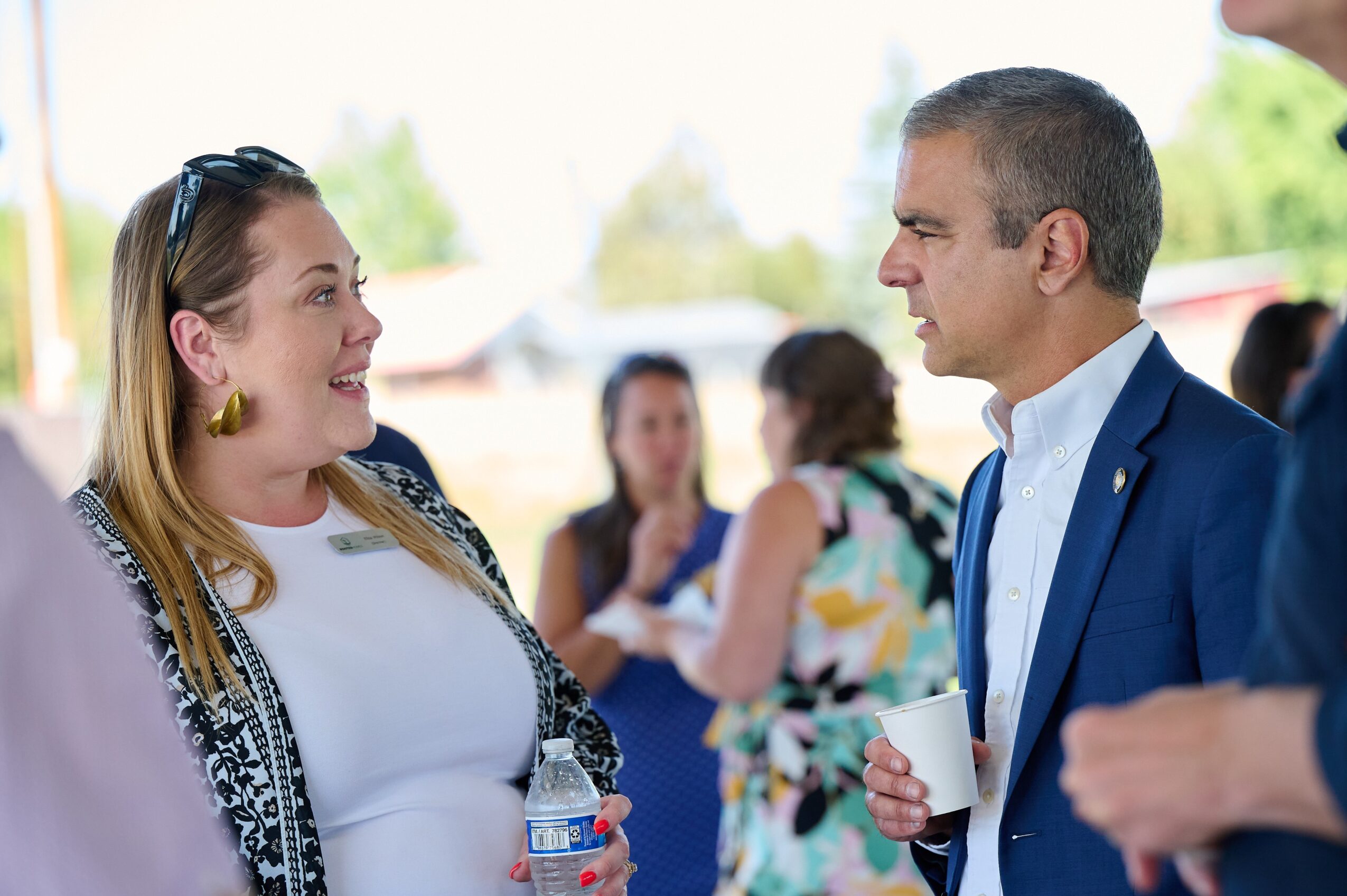 Two people engaged in conversation outdoors, with other people in the background, some holding cups, in a bright setting.