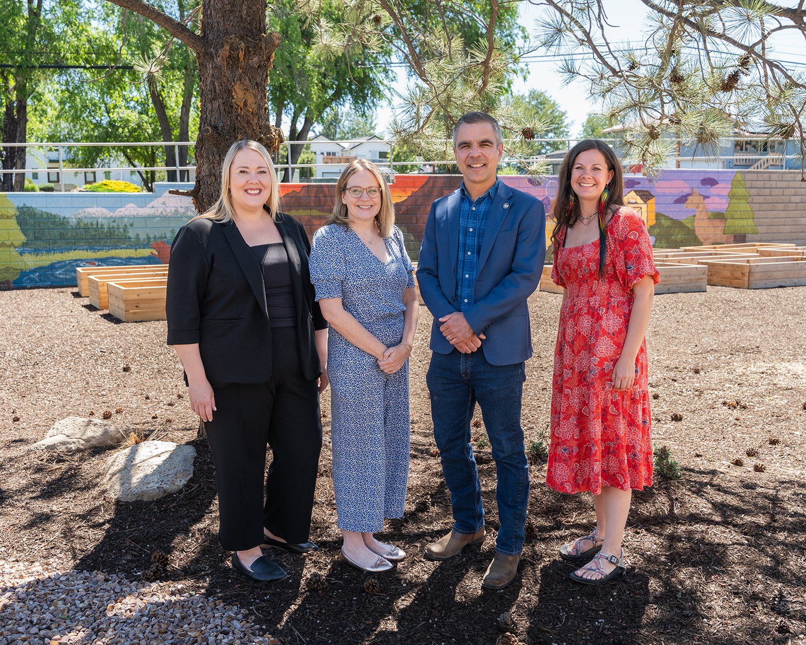 Four people stand outdoors on a sunny day, smiling, with trees and a mural in the background.