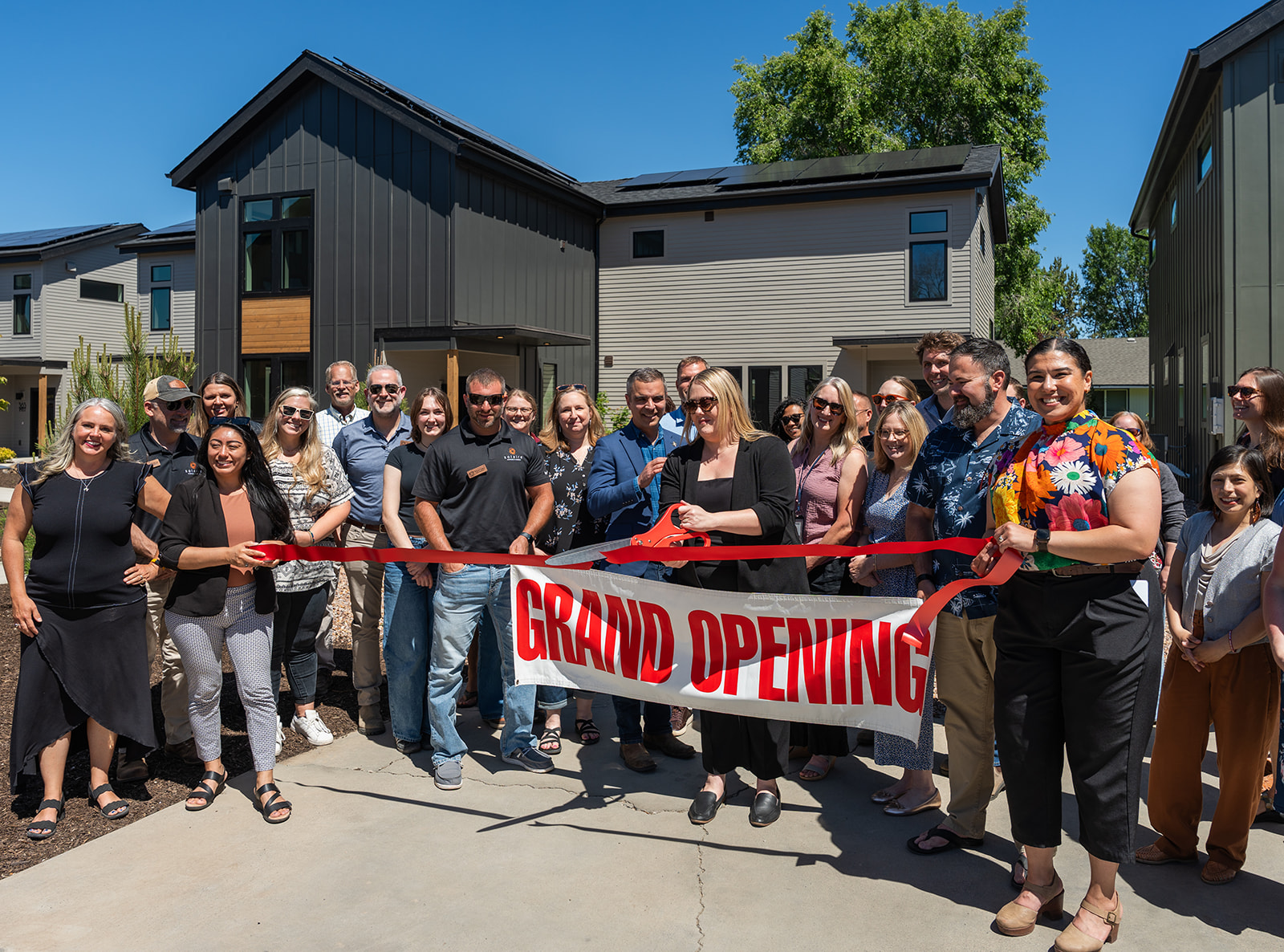 Group of people at a grand opening event holding a red ribbon outside modern buildings, with trees and blue sky in background.