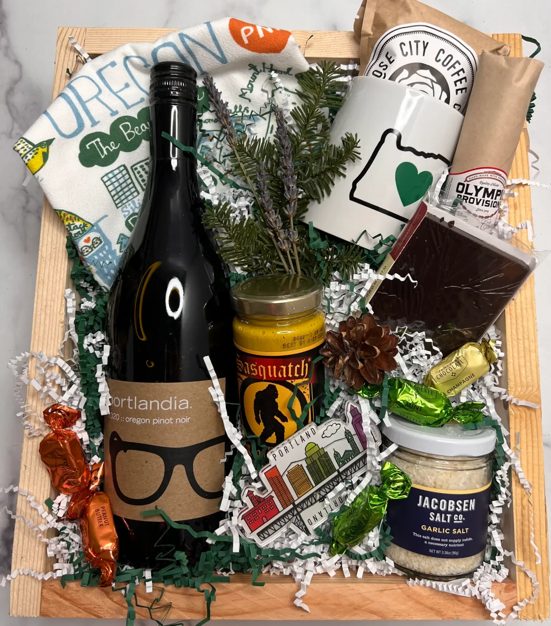 Gift basket with a bottle, jar, coffee, snacks, and greenery, decorated with candies and paper shreds.