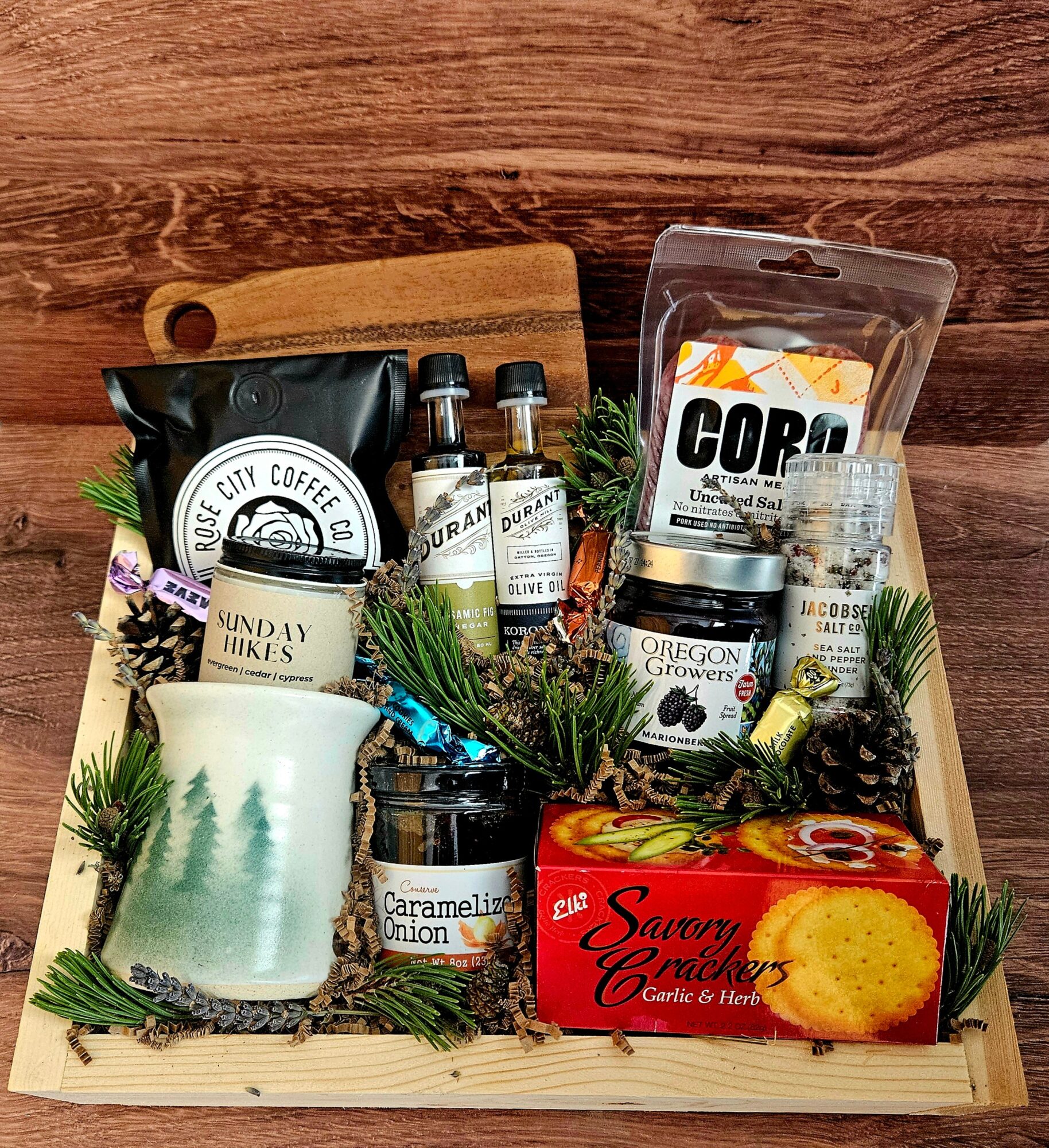 Wooden tray with coffee, tea, and snack items, including a candle, small bottles, a box of cookies, and greenery.