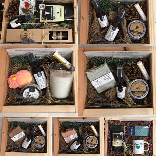 Six wooden boxes filled with skincare, candles, and small gifts, decorated with greenery and pinecones, arranged in two rows.