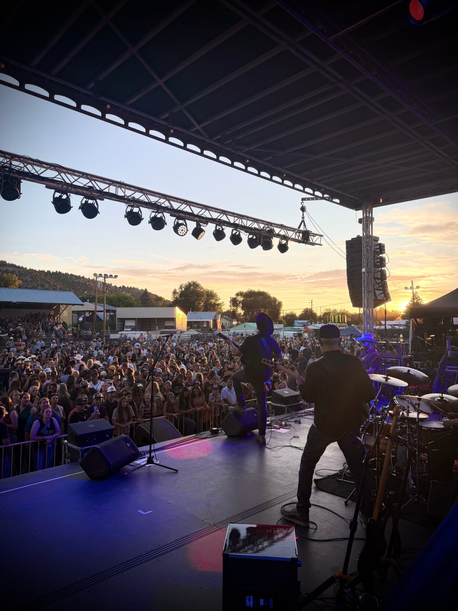 Concert stage with musicians performing outdoors, audience watching, sunset sky, and stage equipment visible.
