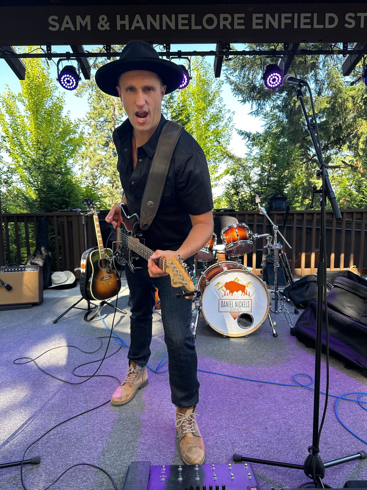 Musician playing guitar on stage outdoors with trees and a fence in the background.
