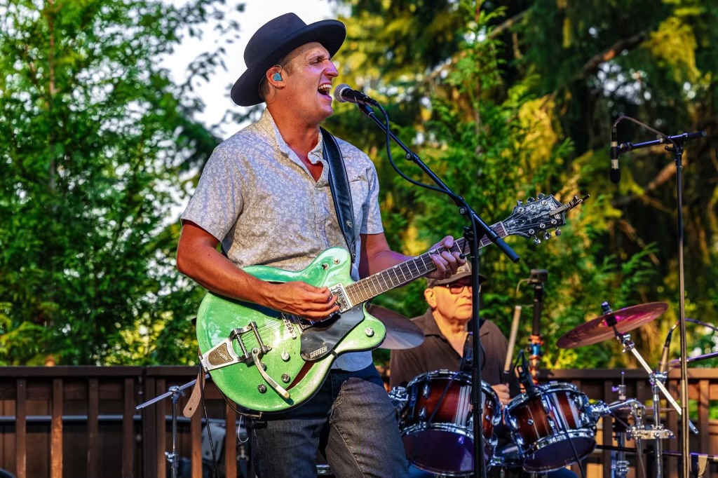Person singing and playing guitar outdoors with trees in background, wearing a hat and patterned shirt.