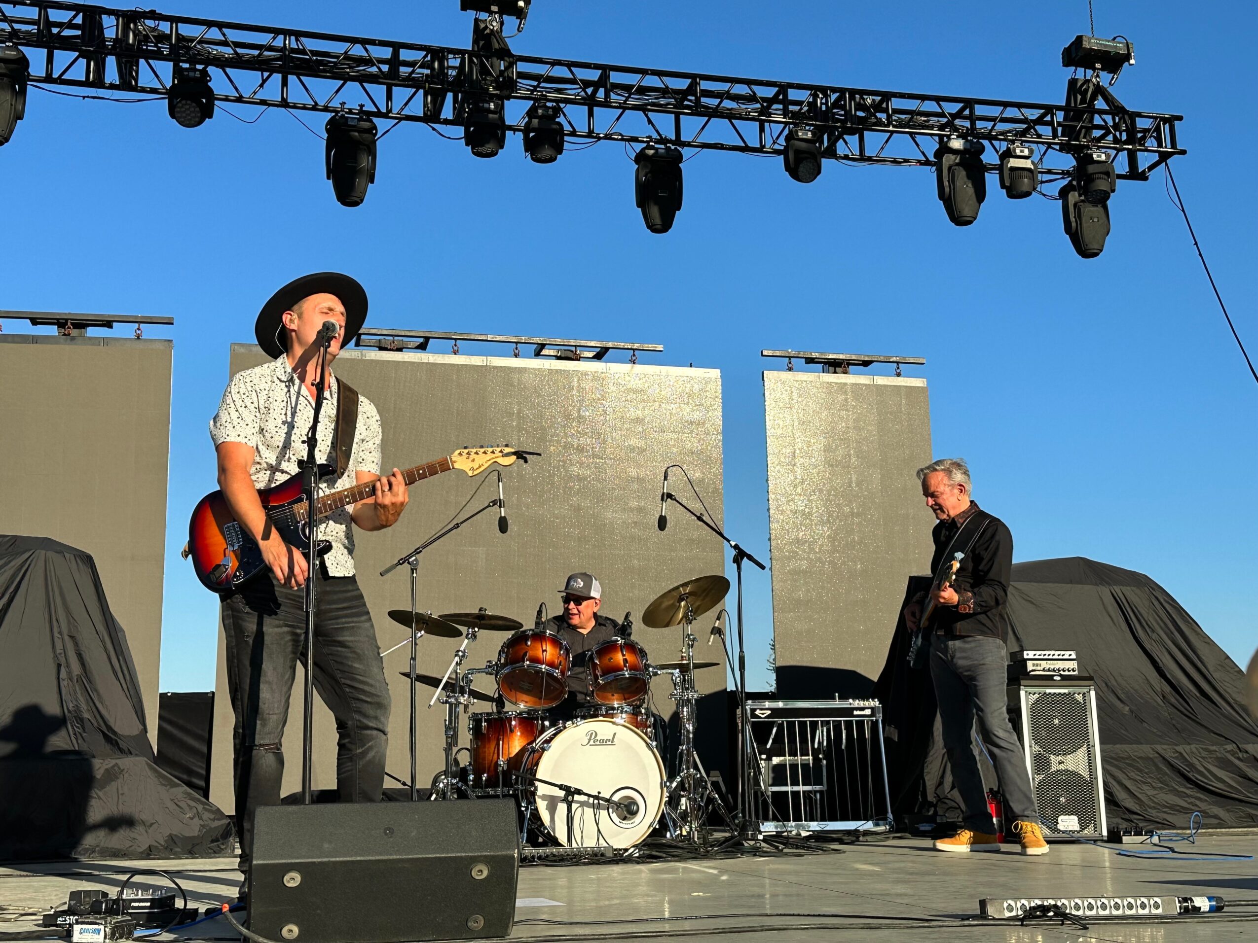Three musicians perform on an outdoor stage with a blue sky, one playing guitar and singing, another on drums, and a third on keyboard.