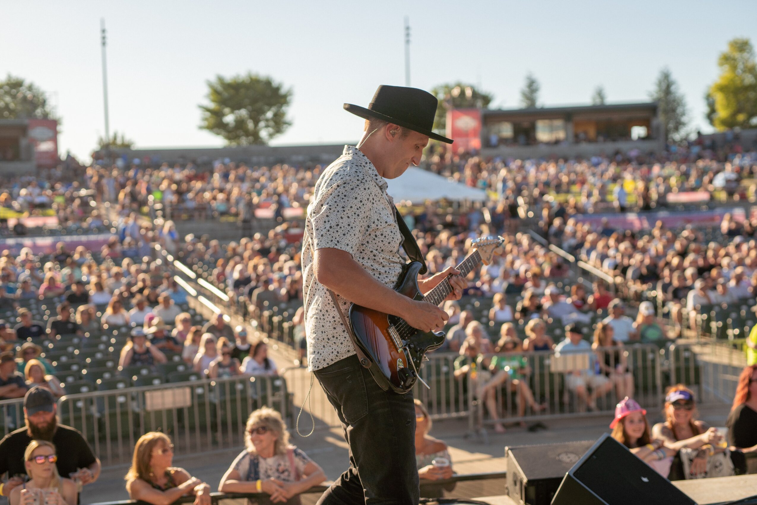 Musician playing guitar on stage at outdoor concert with audience in background.
