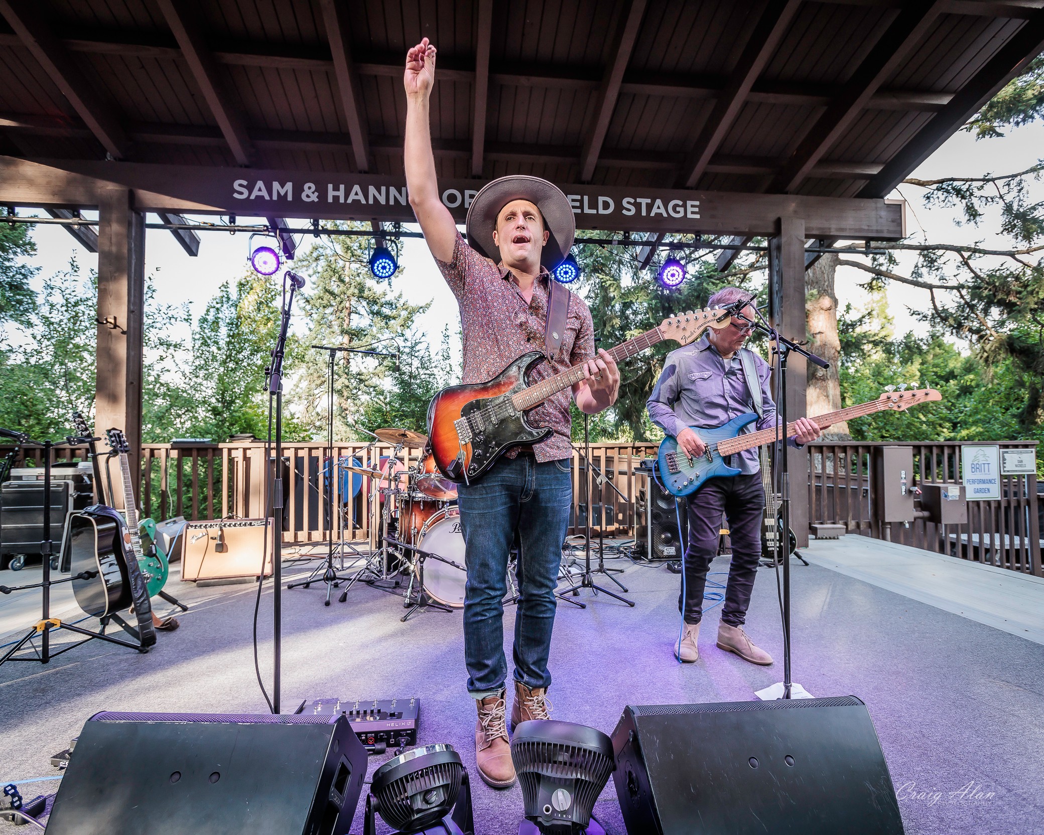 Musicians perform on an outdoor stage with trees in the background, one raising an arm, others playing guitars.