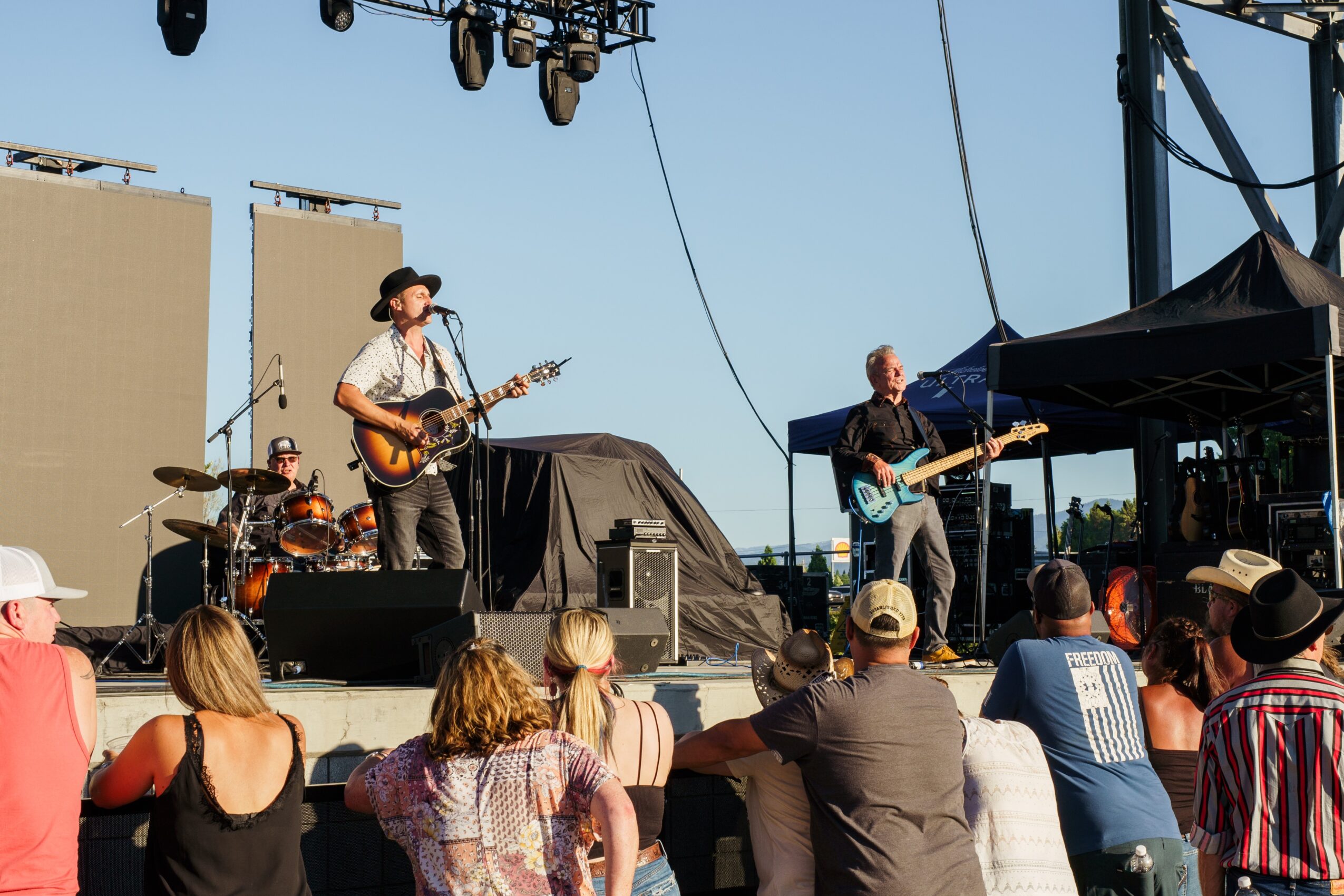 Band performs on stage outdoors with audience watching, some wearing hats, during daytime.