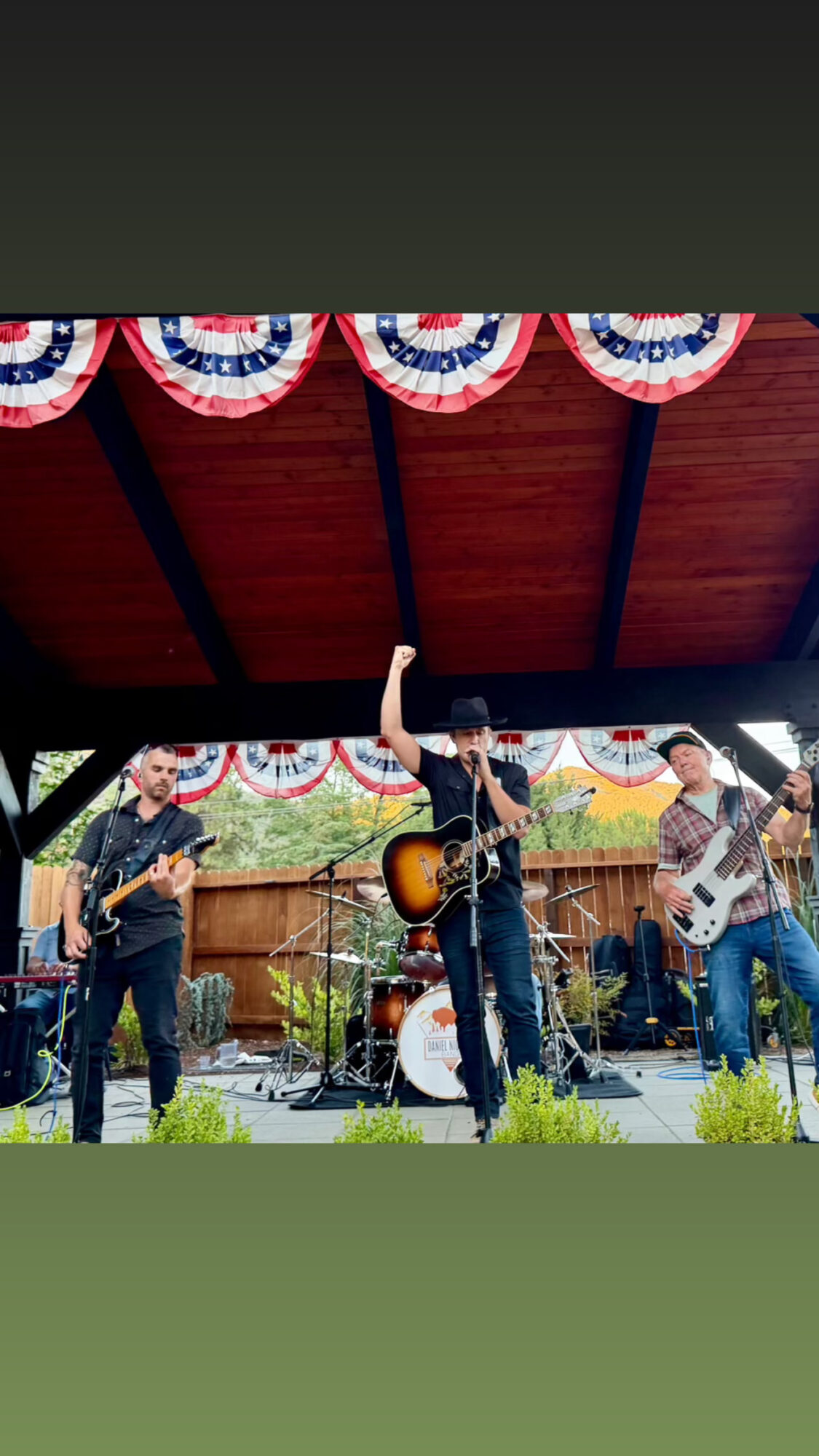 Three musicians perform on an outdoor stage with patriotic bunting, one playing guitar and singing, others playing bass and guitar.