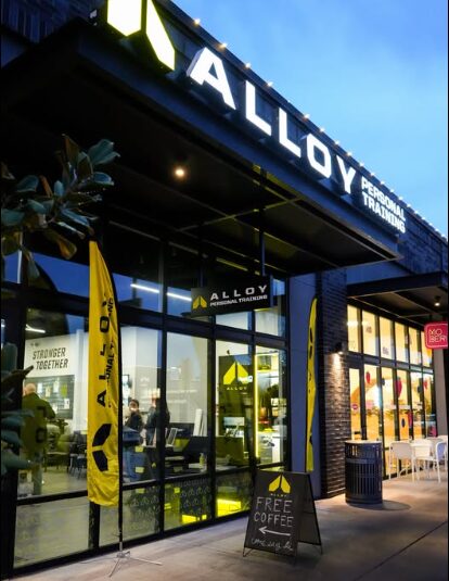 Storefront with large glass windows, yellow banners, and a sign for free coffee outside, under a black awning with illuminated sign.