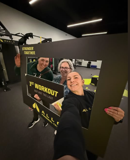Group of four people smiling behind a large frame at the gym, holding a sign that says '1st Workout'.