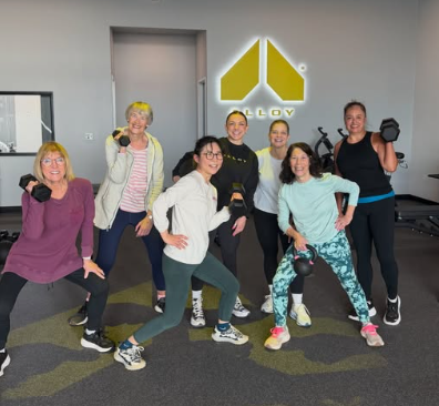 Group of seven women in workout clothes holding dumbbells in a gym with a logo on the wall.