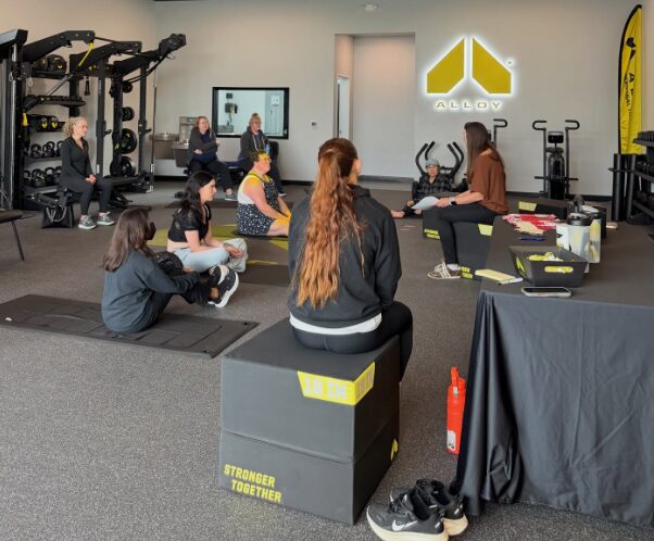 Group of people sitting on mats and chairs in a gym, facing a screen, with fitness equipment around them.