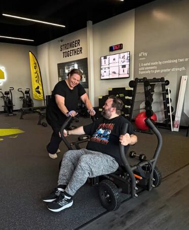 Two people smiling and interacting in a gym, one in a wheelchair and the other standing, with gym equipment in background.