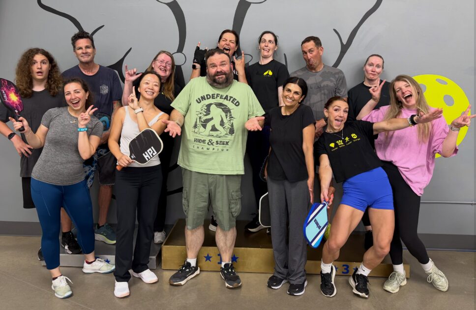 Group of 12 people standing together indoors, some smiling and making gestures, with a painted wall background featuring trees and a yellow shape.