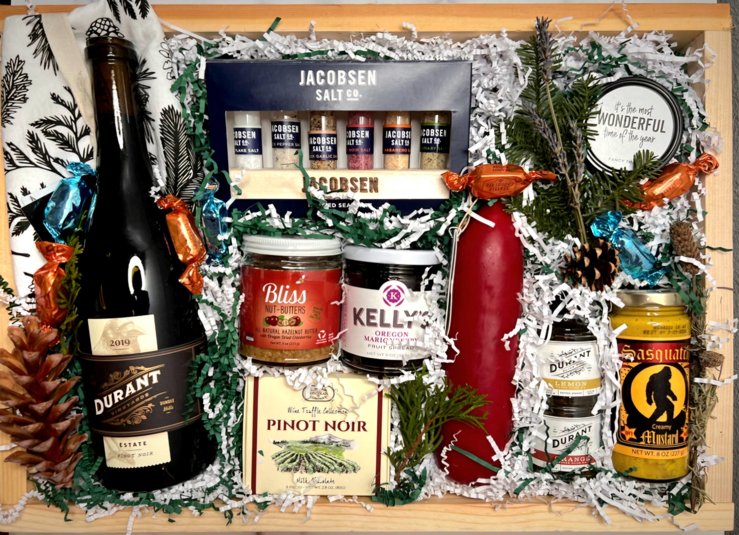 Gift basket with wine, skincare, candles, and decorative pinecones, surrounded by festive greenery and white shredded paper.