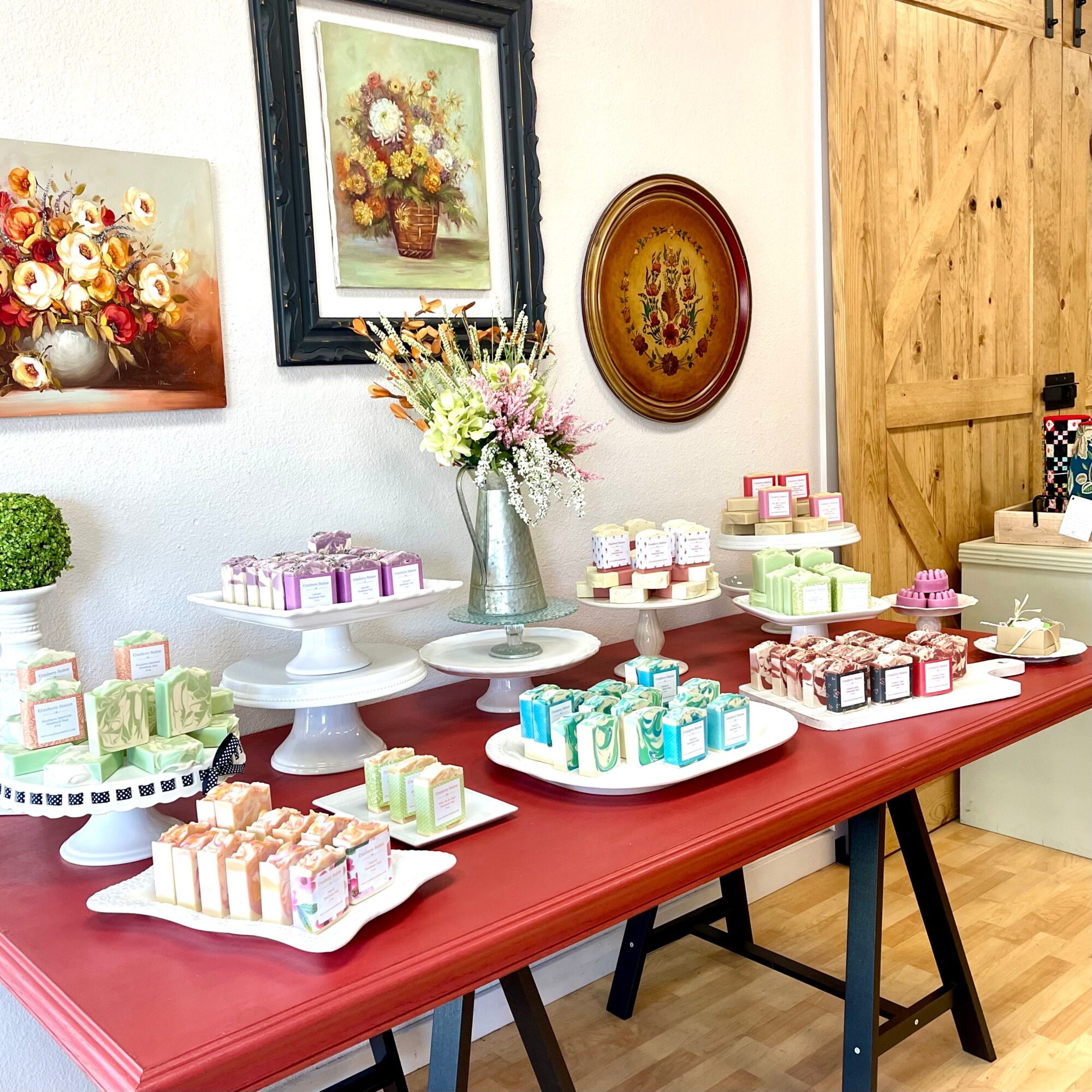 Table with colorful soap bars and floral arrangements, decorated with framed paintings and a wooden wall in the background.