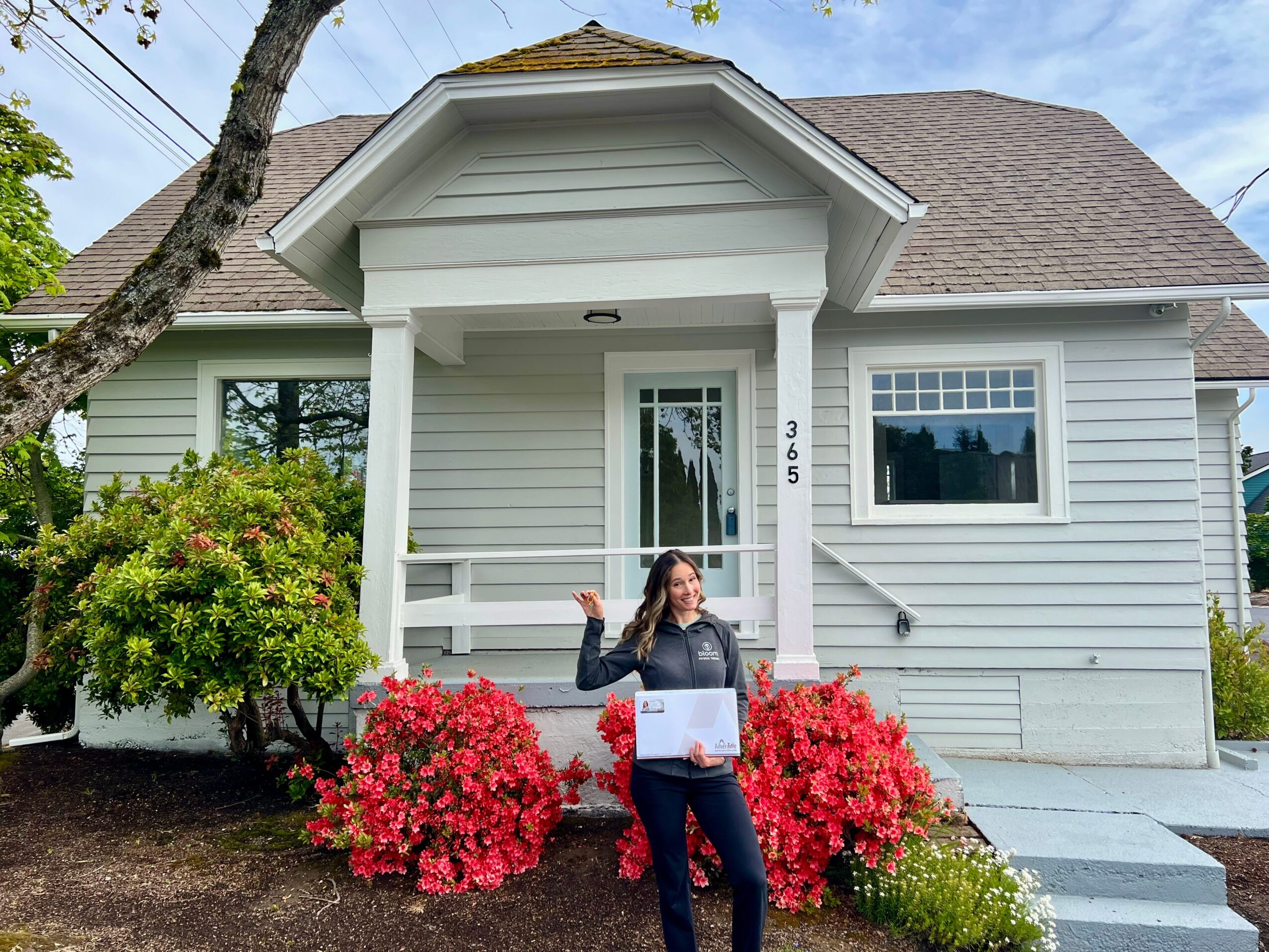 Woman standing in front of a house with white siding, holding a box, surrounded by pink bushes, with a tree on the left.