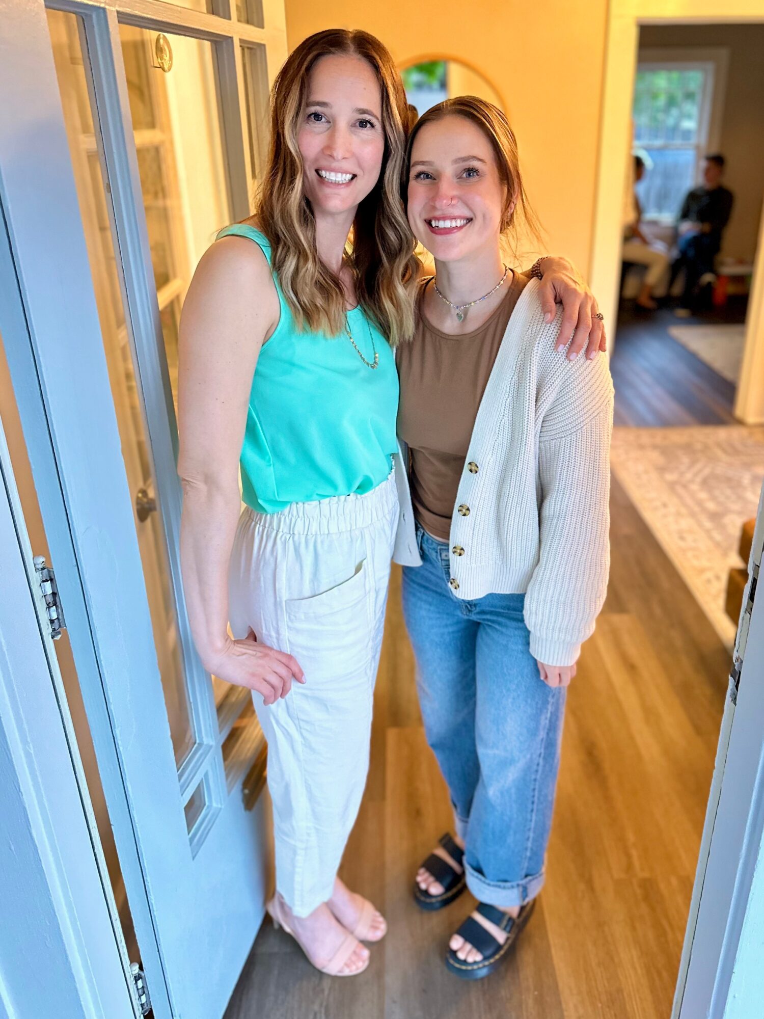 Two women standing indoors, smiling, with one arm around each other, in a brightly lit room with yellow walls.