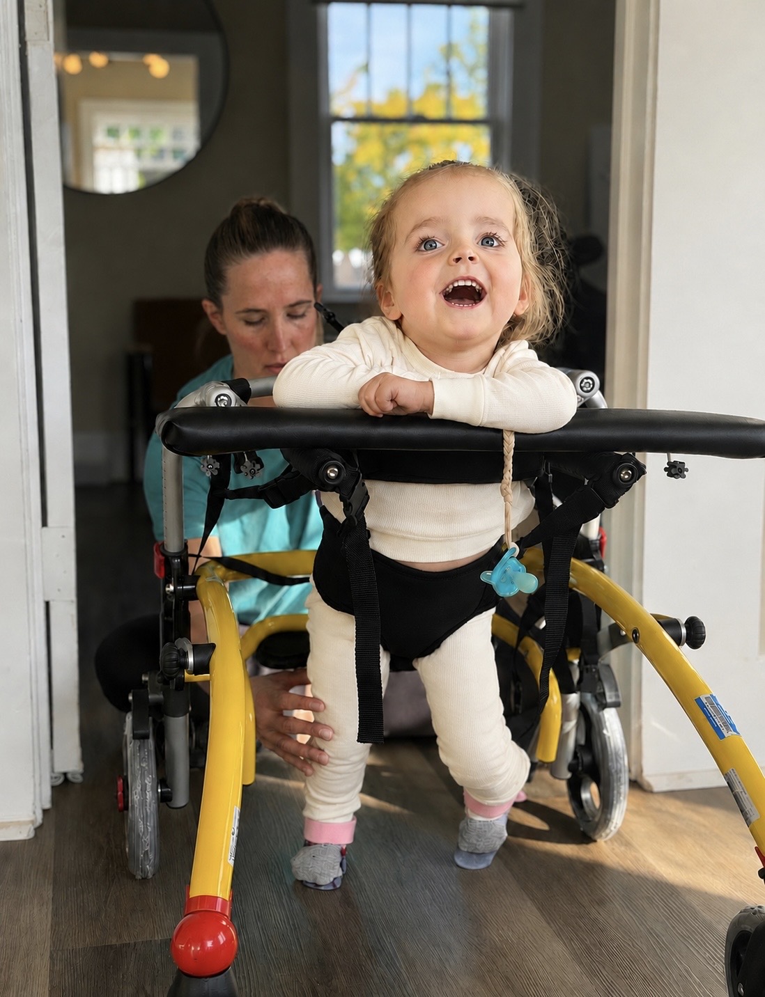 Child in a walker smiling with a woman behind, in a room with large windows and a mirror, sunlight outside.