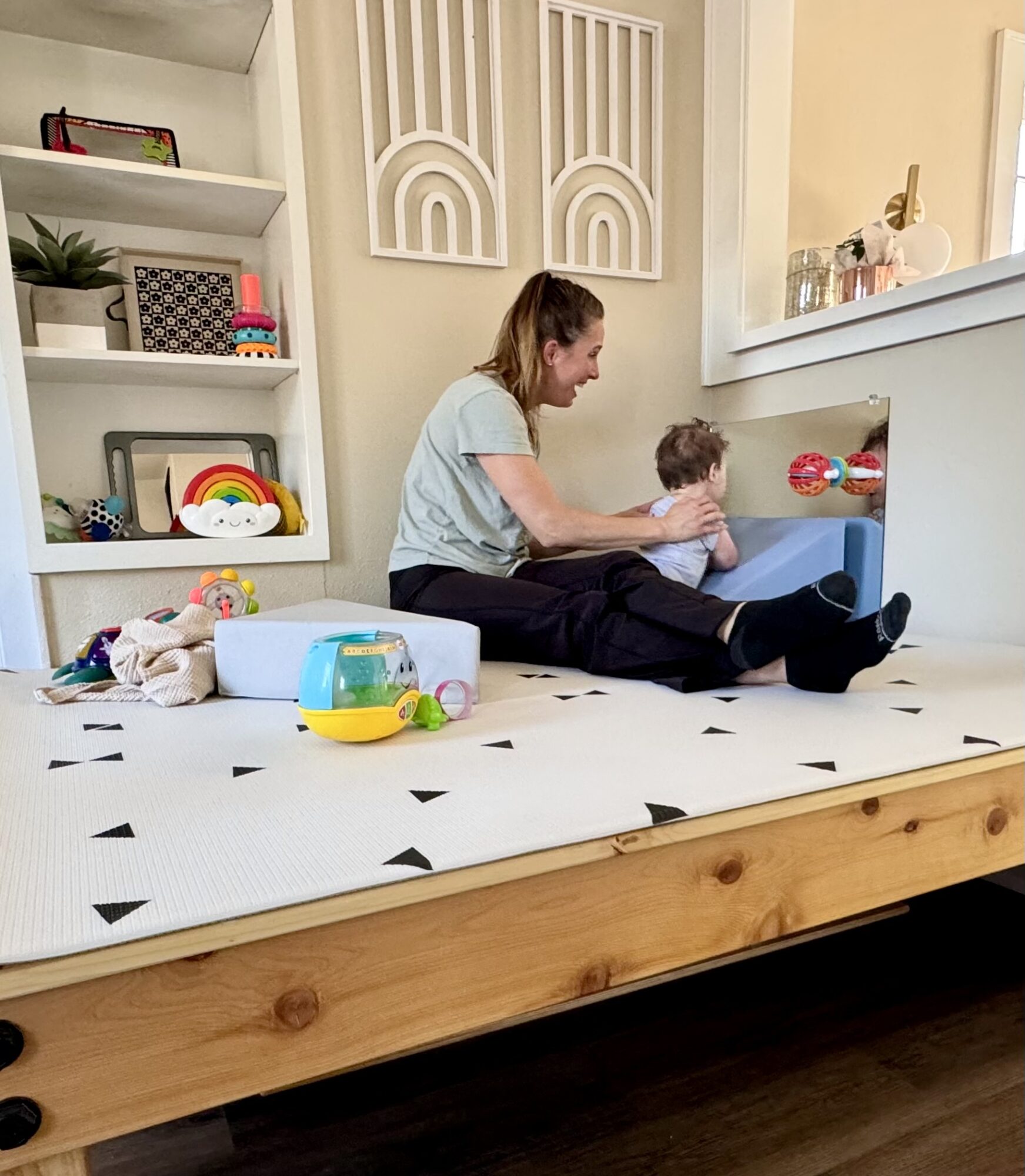 Woman and child playing with toys on a white and black patterned mat in a bright room with shelves and decorations