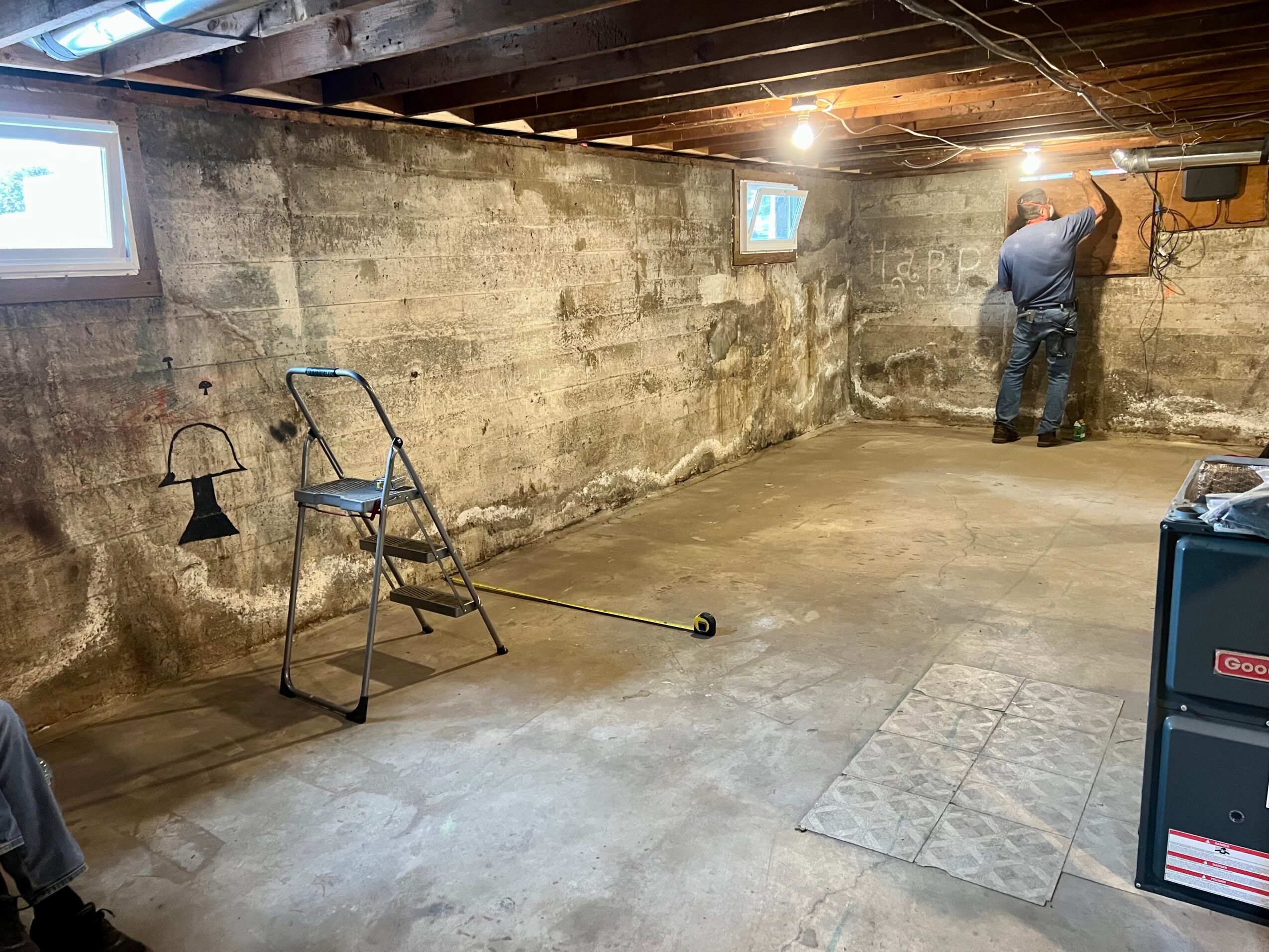 Person working on wall in basement with concrete walls and ceiling, tools and a chair nearby, small windows, and a floor mat.