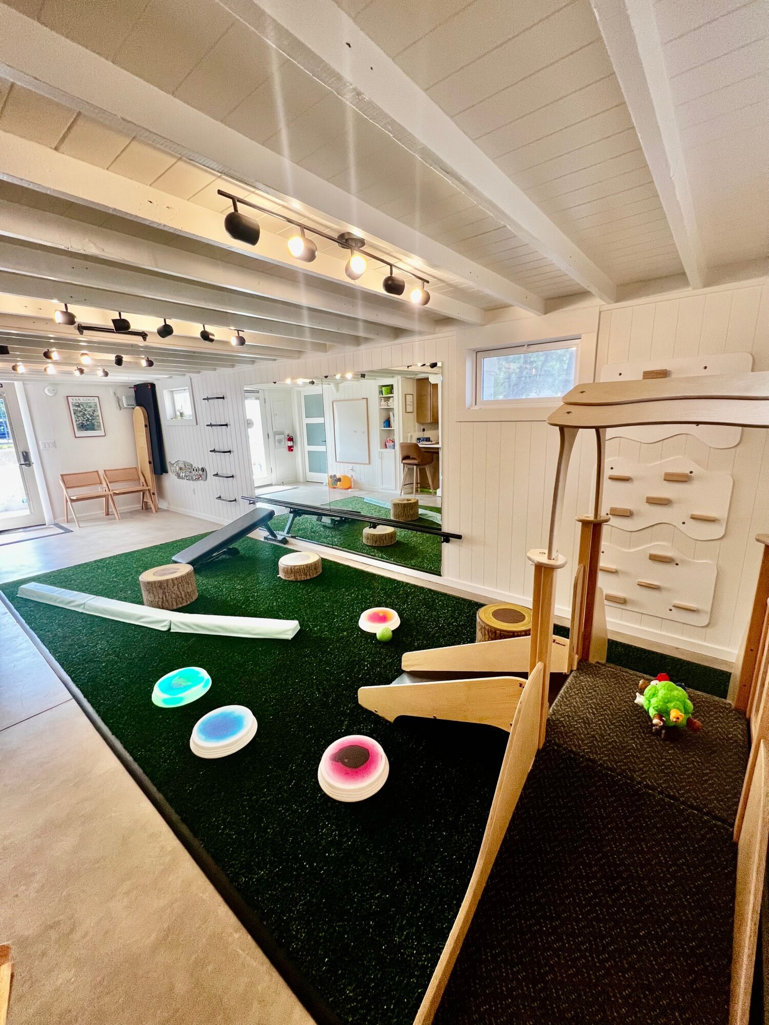 Indoor play area with a green carpet, wooden play structures, and colorful circular objects scattered on the floor.
