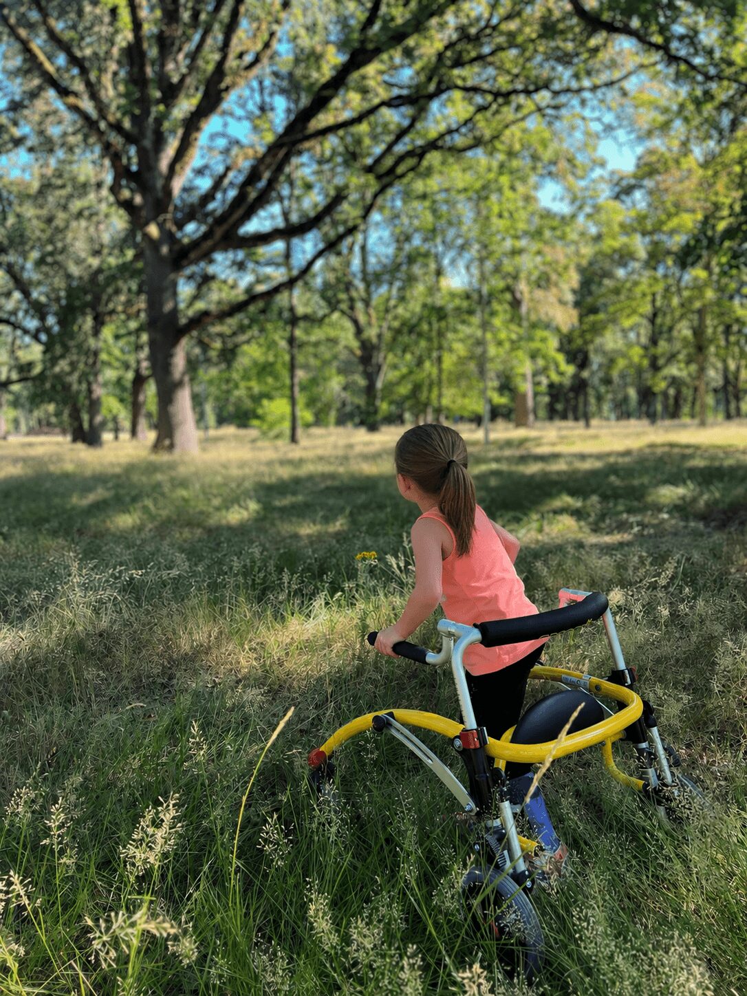 Girl in pink shirt standing next to a yellow and white bicycle in a grassy park with trees and blue sky.