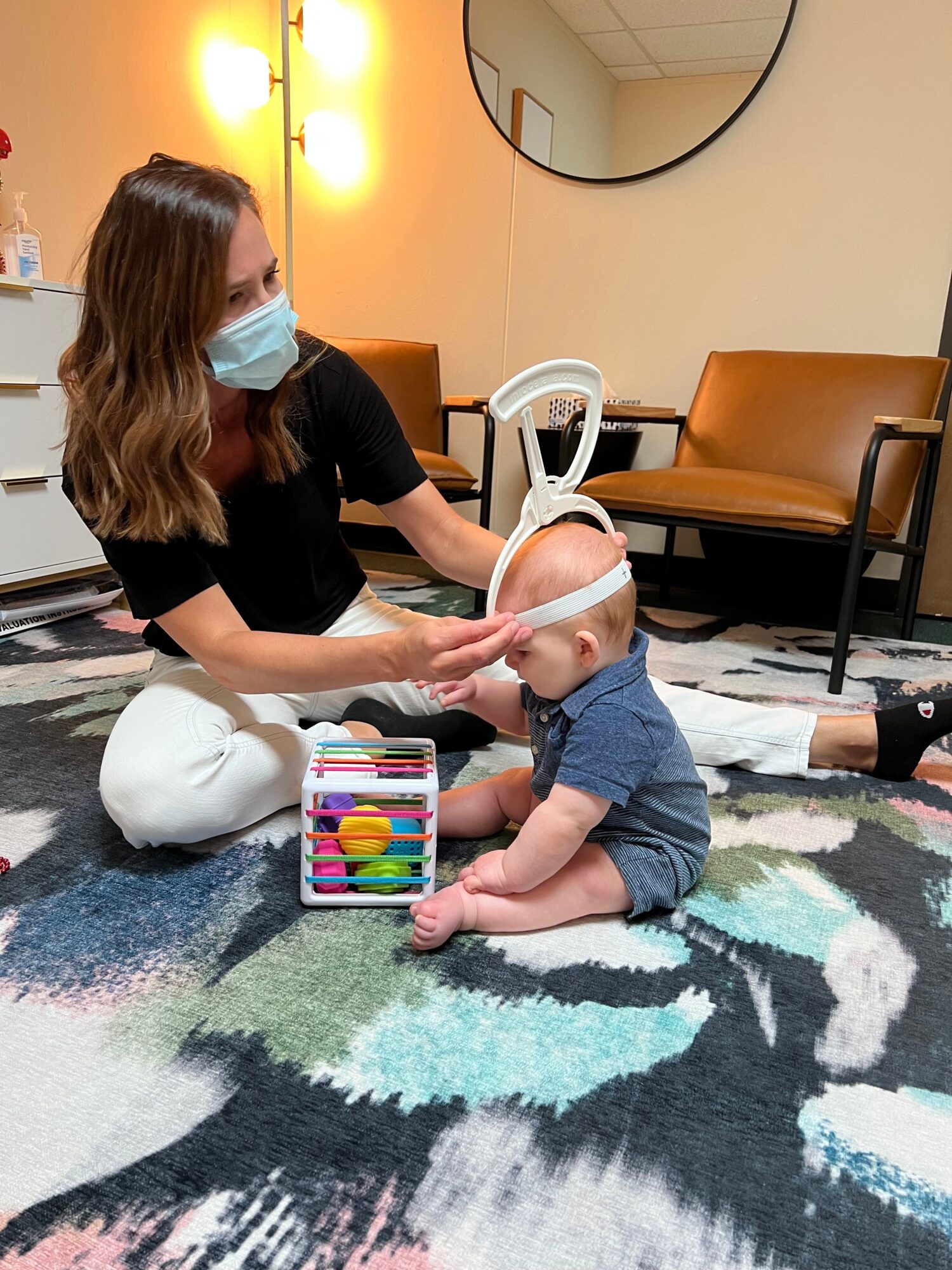 Adult and child sitting on a colorful rug, playing with a toy tablet, both wearing face masks.