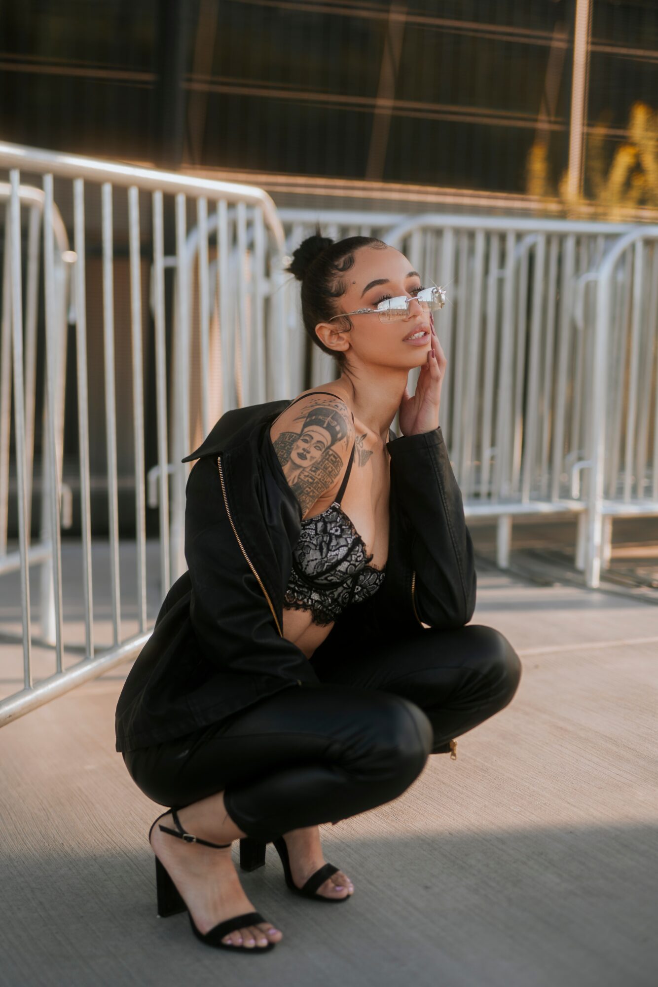 Woman crouching with glasses, tattoos, and black outfit, outdoors near metal barriers, looking thoughtful.