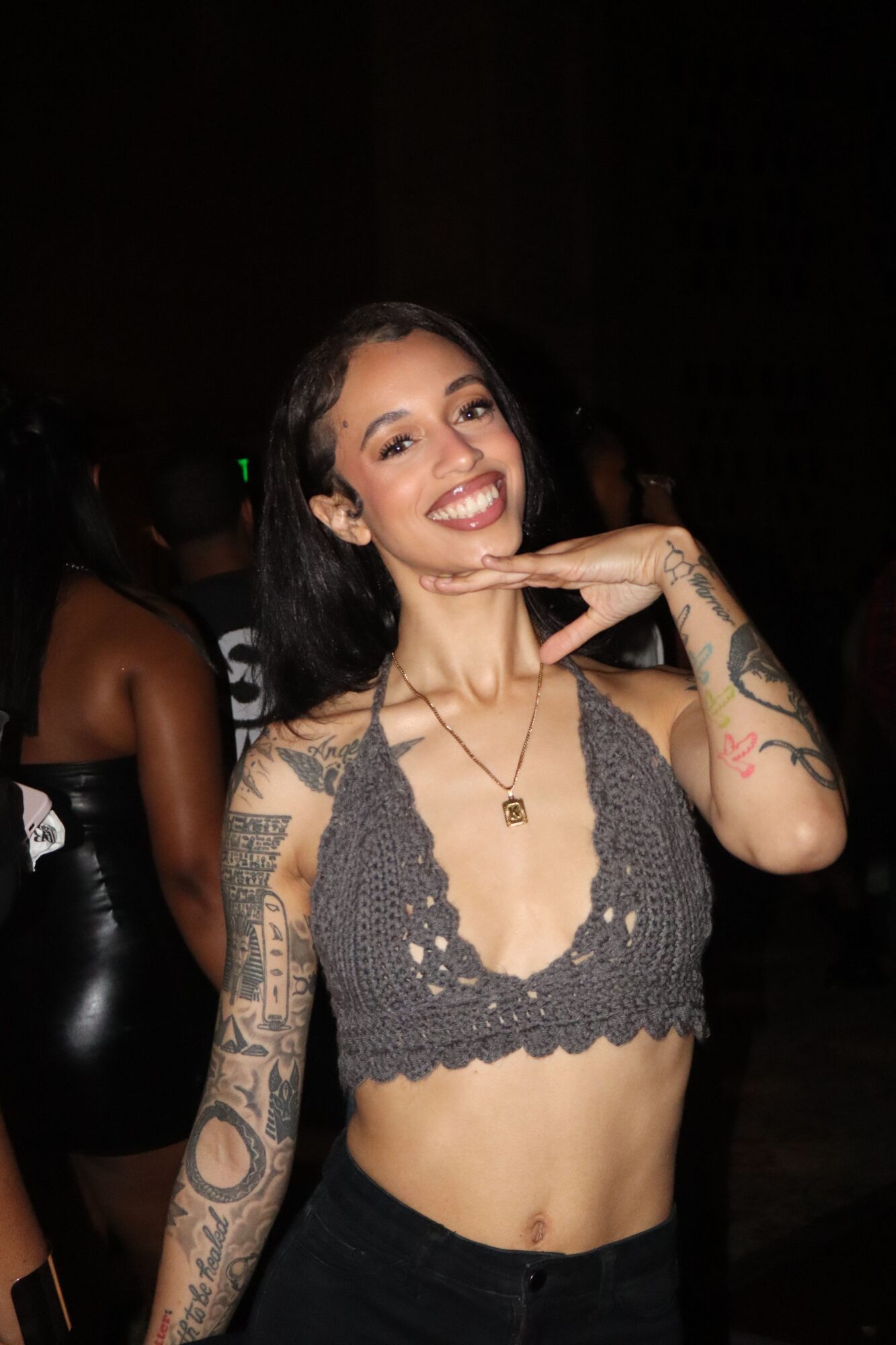 Woman with tattoos, wearing a gray crochet top, smiling and making a peace sign gesture, dark background.