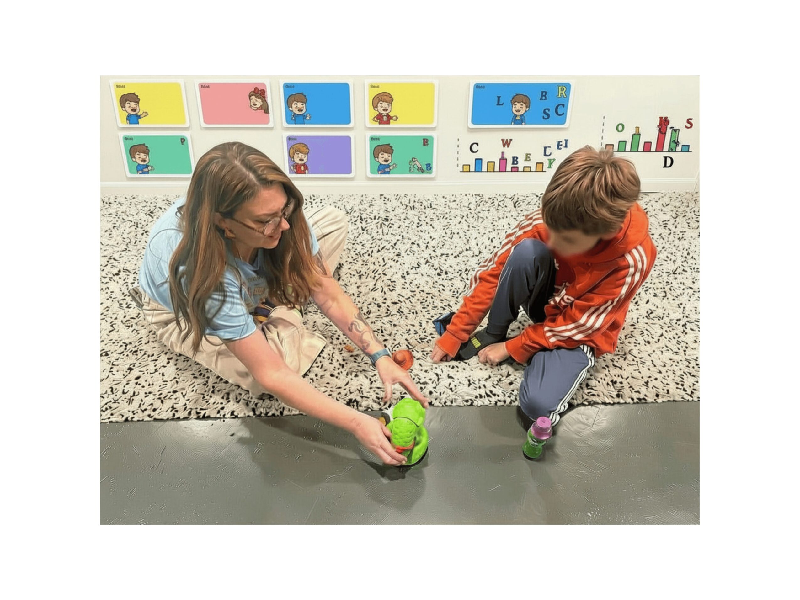Two children sit on a speckled floor, playing with a green ball, with colorful educational posters behind them.