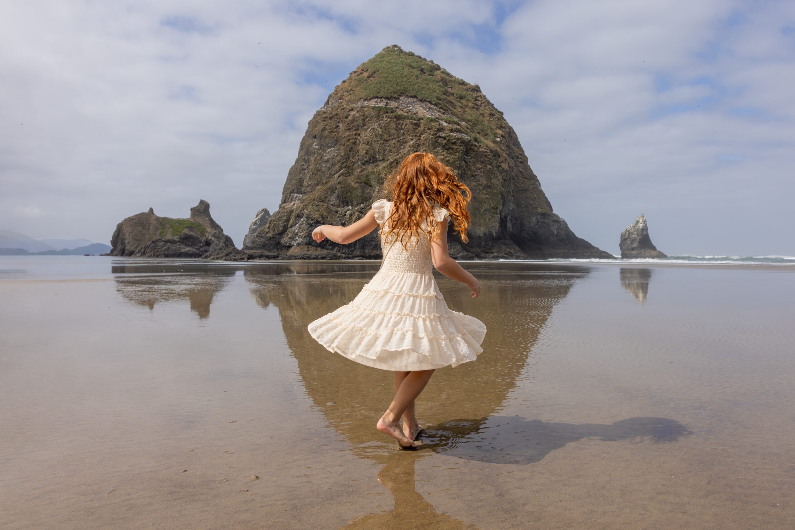 Red-haired girl in a white dress twirling on wet sand with large rock formations in the background, reflection visible in water.