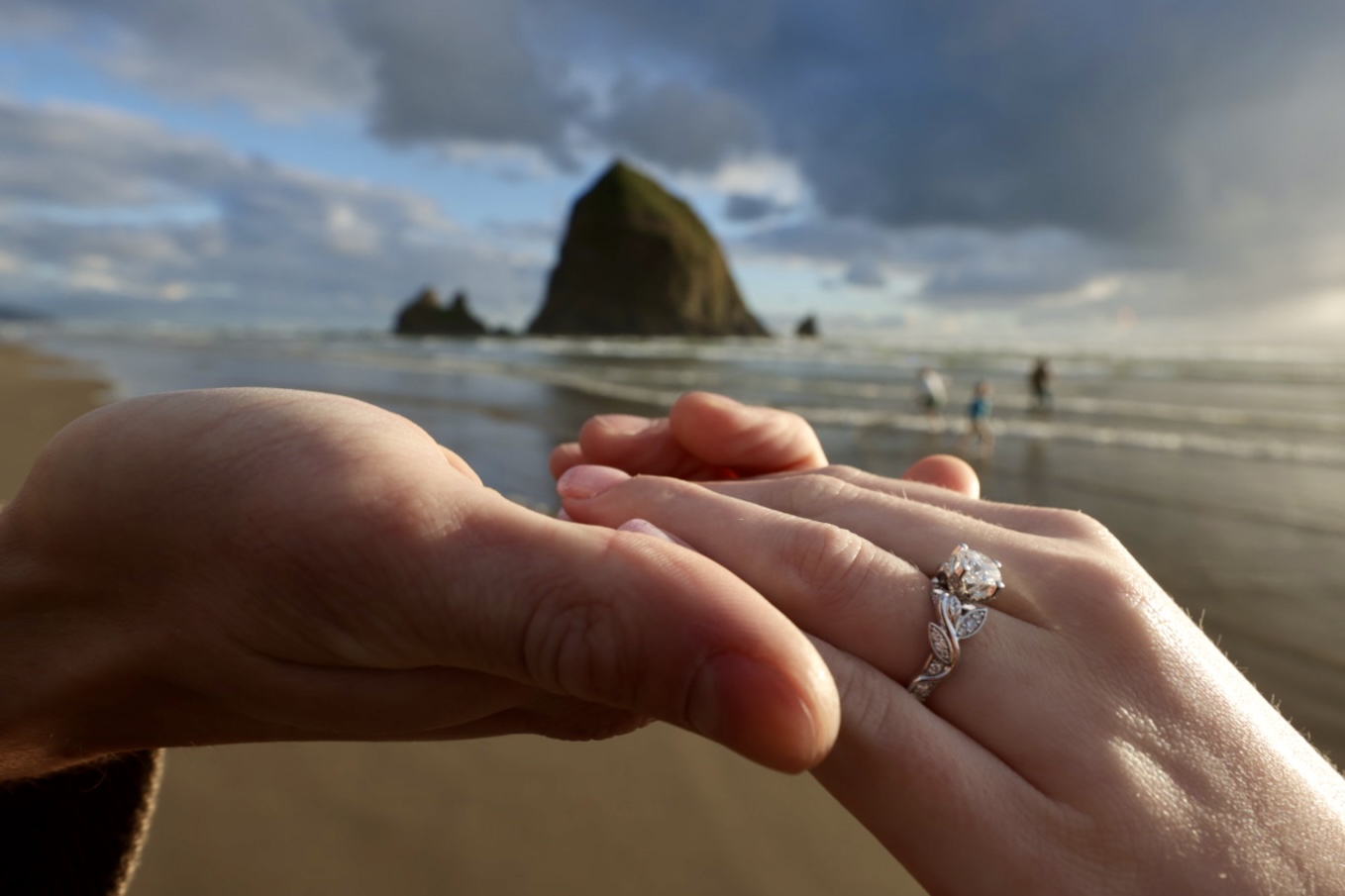 Two hands with a ring on one finger, held near the beach with a large rock formation and cloudy sky in the background.