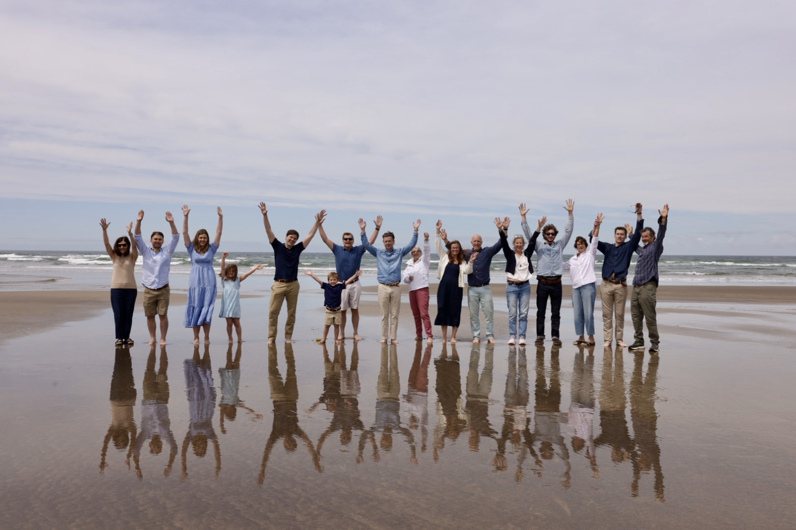 Group of people standing on beach with arms raised, reflected in wet sand, ocean and cloudy sky in background.