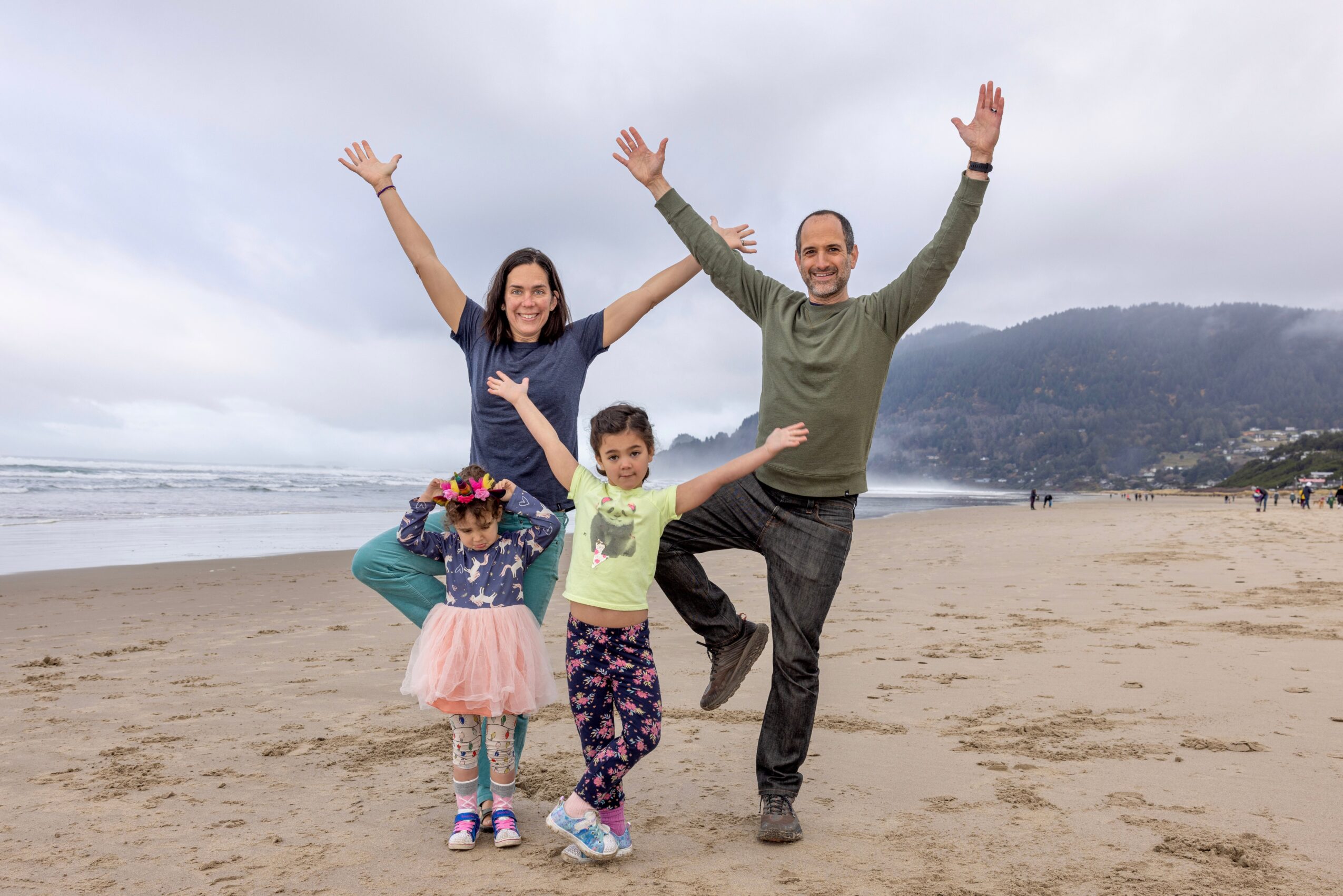 Family of four on a beach, smiling and raising arms, with mountains and ocean in background.