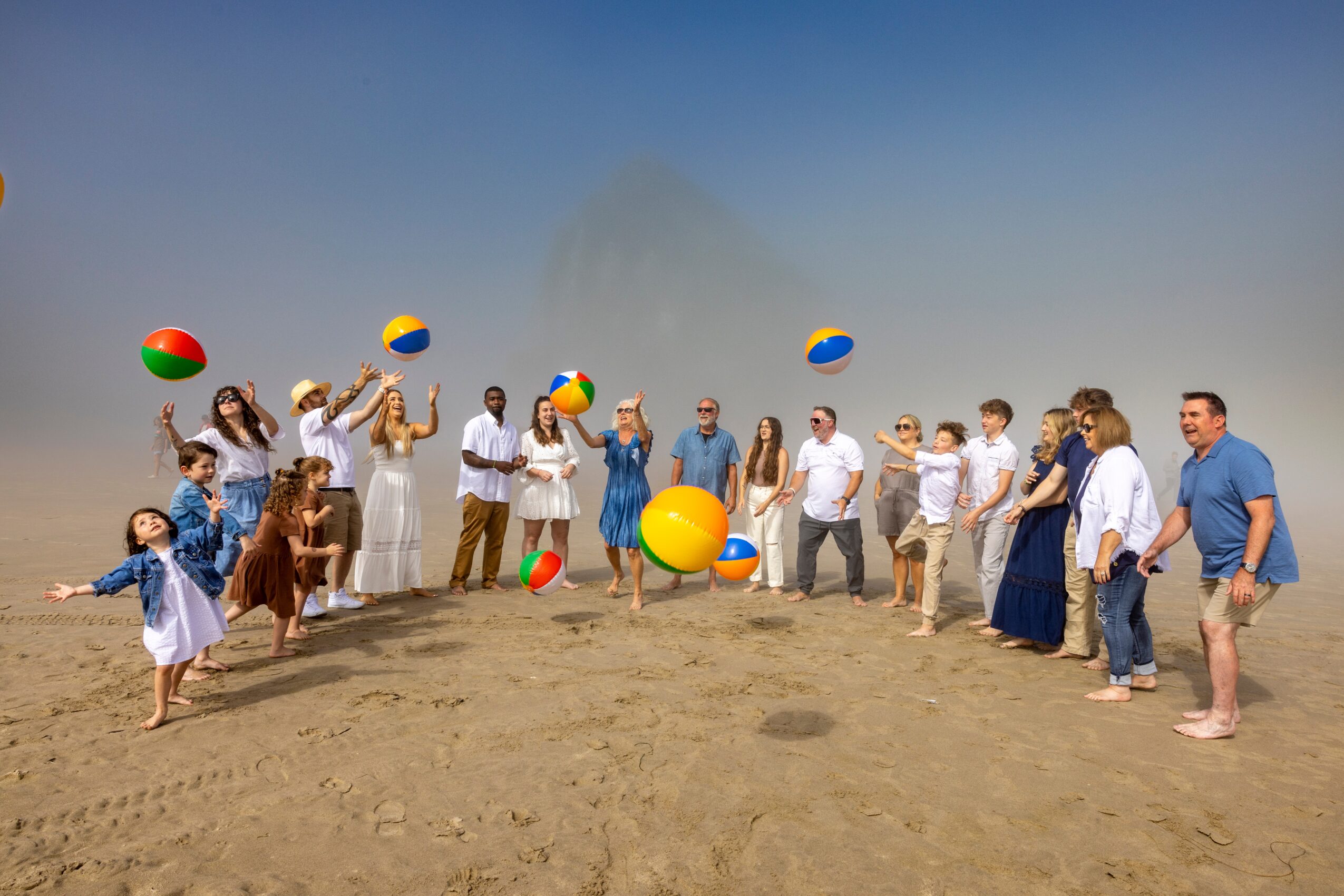 People and children playing with beach balls on a sandy beach under a clear blue sky.