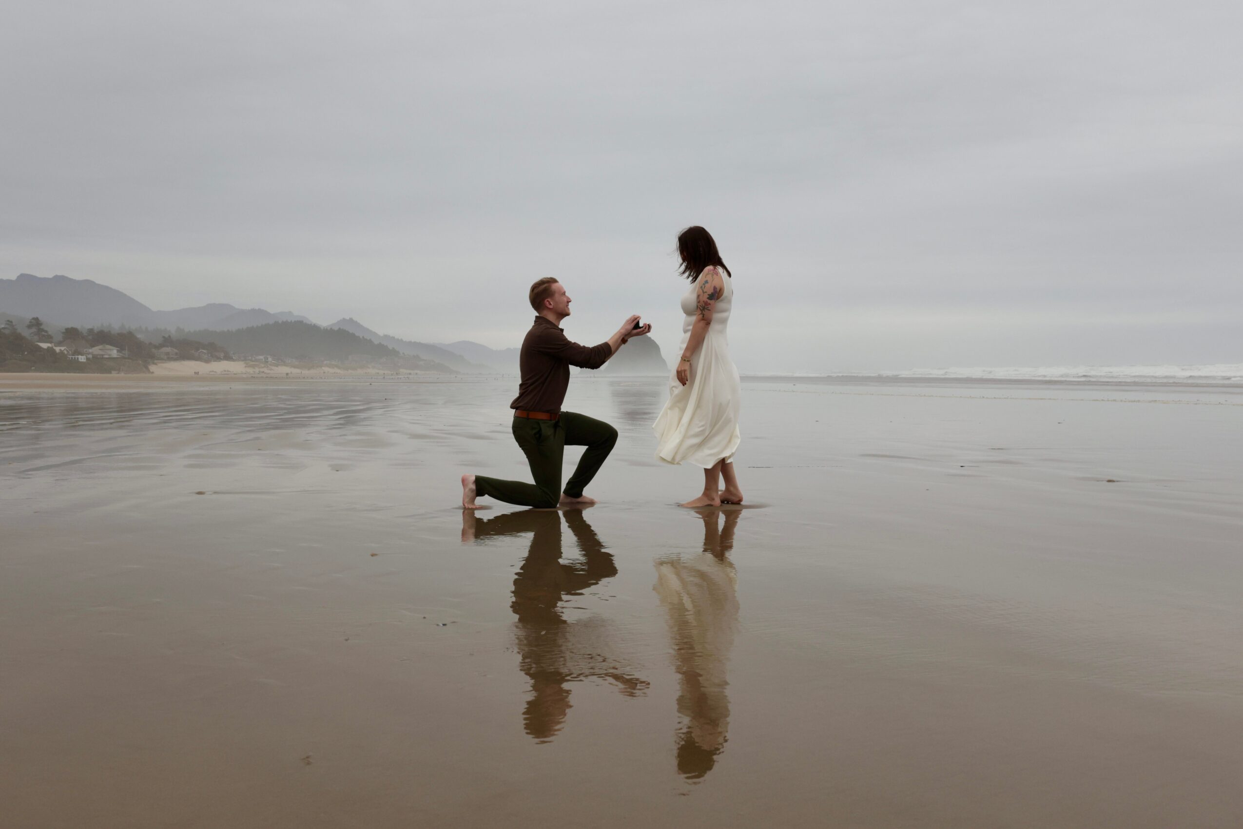 Person kneeling on wet sand proposing to another person standing by the ocean, with a cloudy sky and distant hills in background.