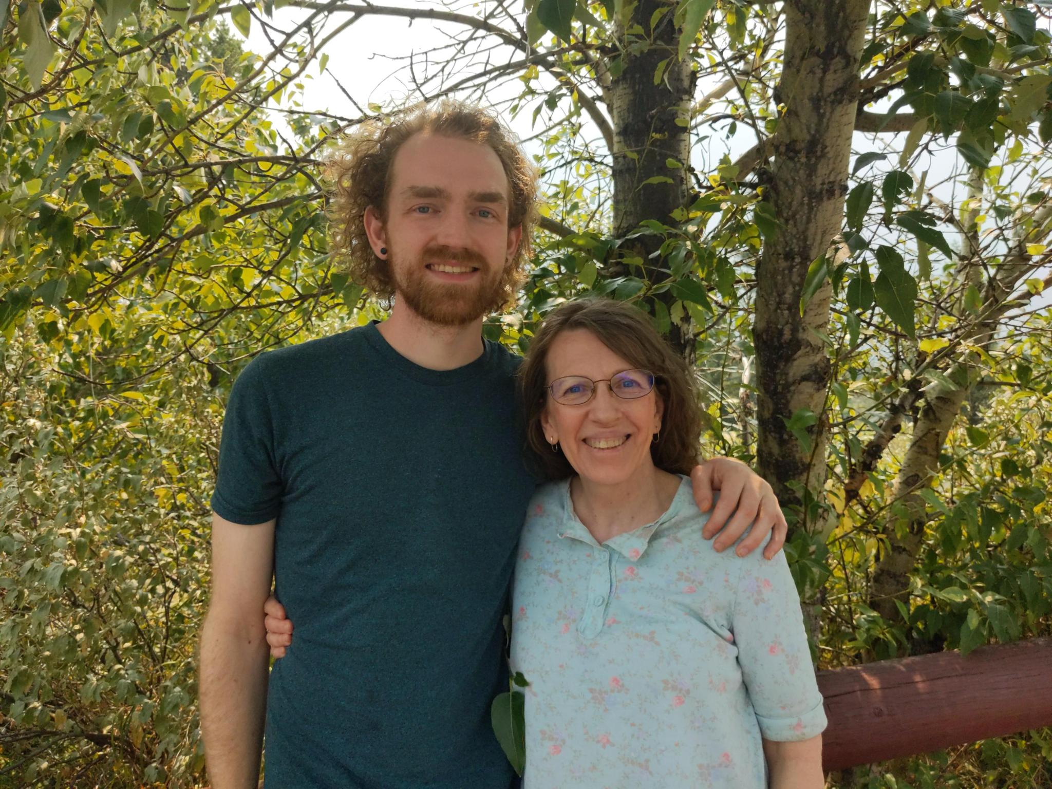A man and woman stand outdoors among trees, smiling, with the man's arm around the woman's shoulder.