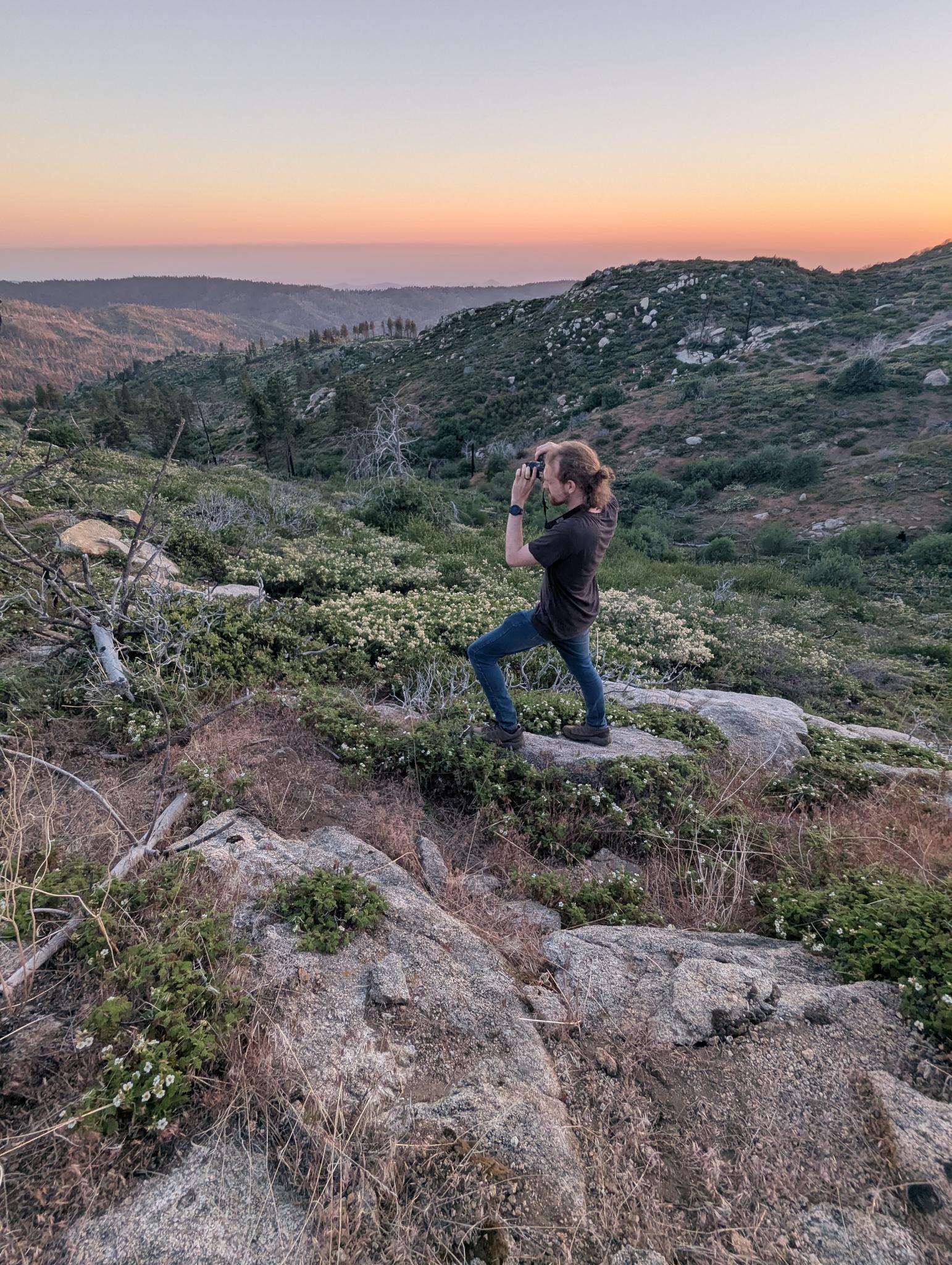 Person standing on rocks in a hilly landscape during sunset, holding a camera or phone, with a scenic view of hills and sky.