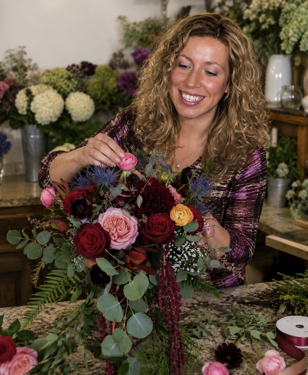 Woman with curly hair smiling while arranging a colorful flower bouquet in a floral shop.