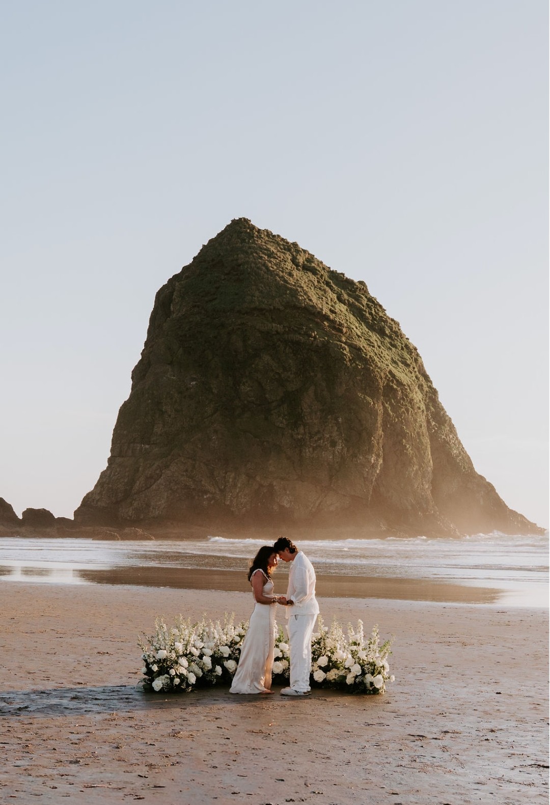A couple in wedding attire kissing on a beach with a large rock formation in the background, surrounded by flowers.