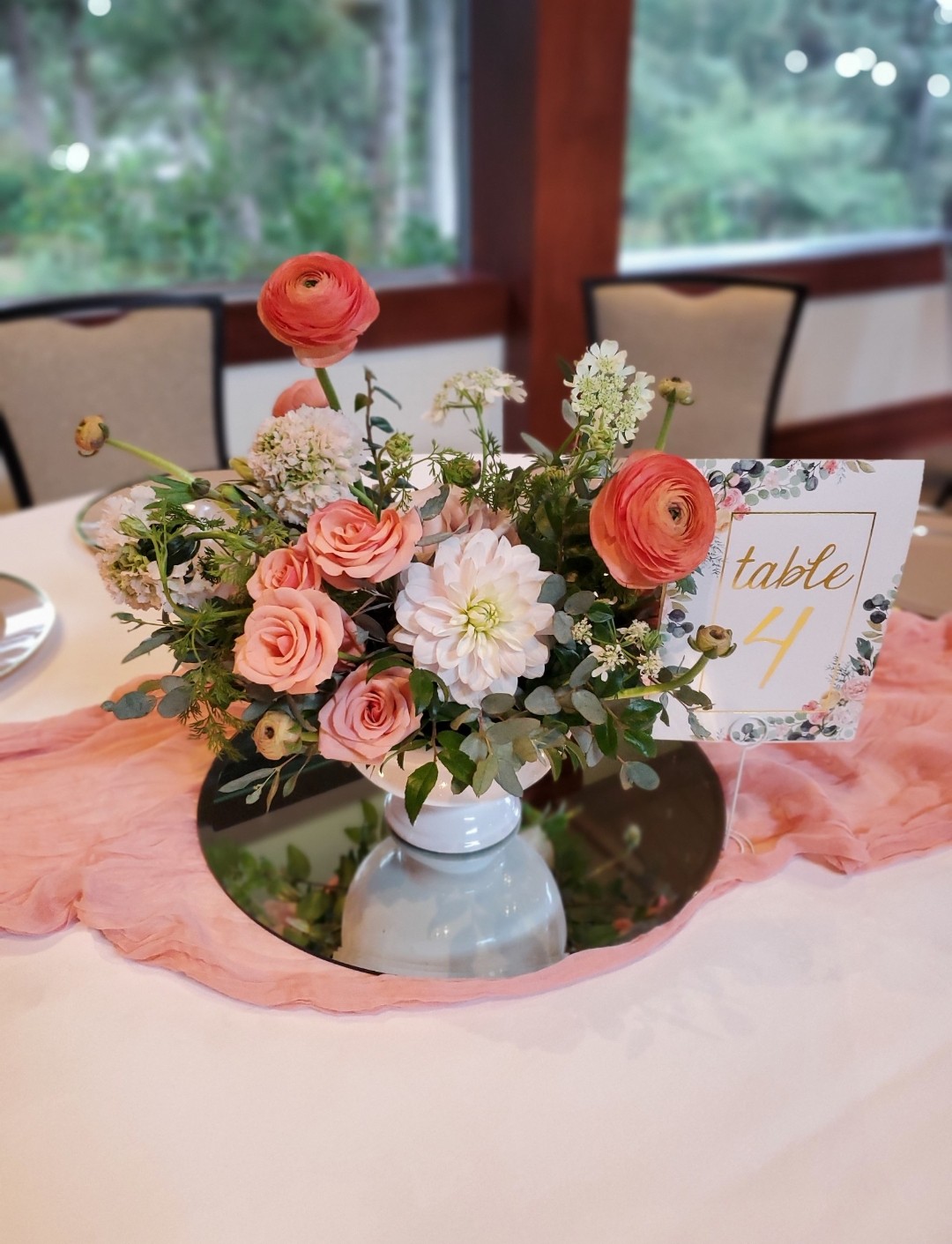 Flower arrangement with pink, white, and orange flowers in a white vase on a table, with a table number card nearby.