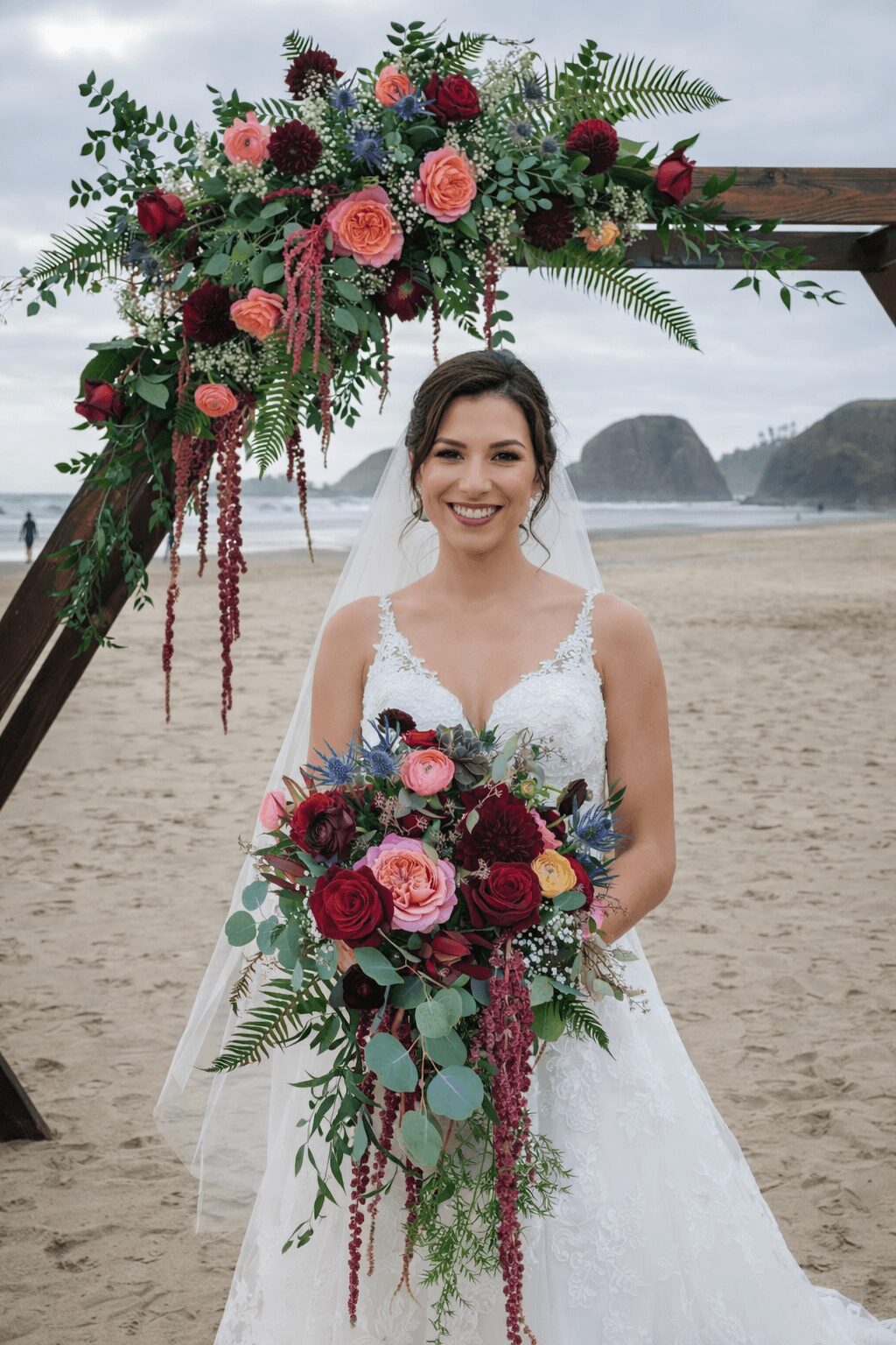 Bride in wedding dress holding a large bouquet, standing under a floral arch on a beach, smiling.